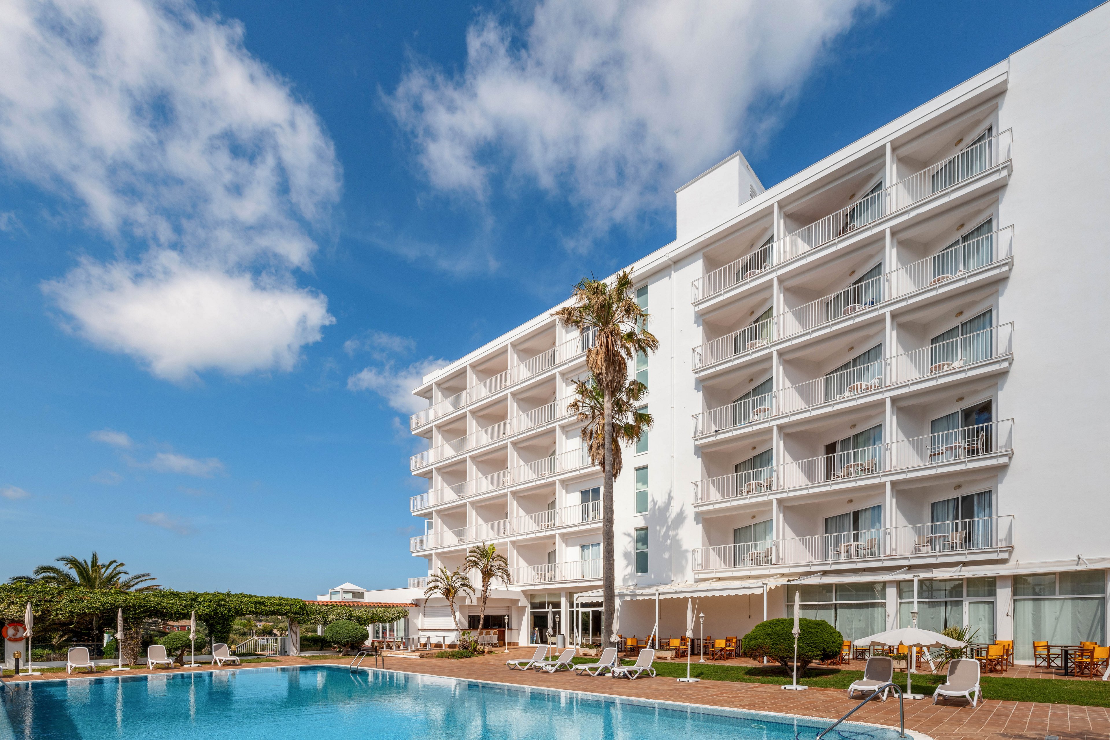 A multi-story white hotel building overlooks a large outdoor swimming pool. Sun loungers and umbrellas are arranged around the poolside terrace, suggesting a relaxing vacation setting. Tall palm trees and manicured greenery frame the scene under a bright blue sky with scattered clouds. The overall atmosphere is calm, sunny, and inviting for resort guests.