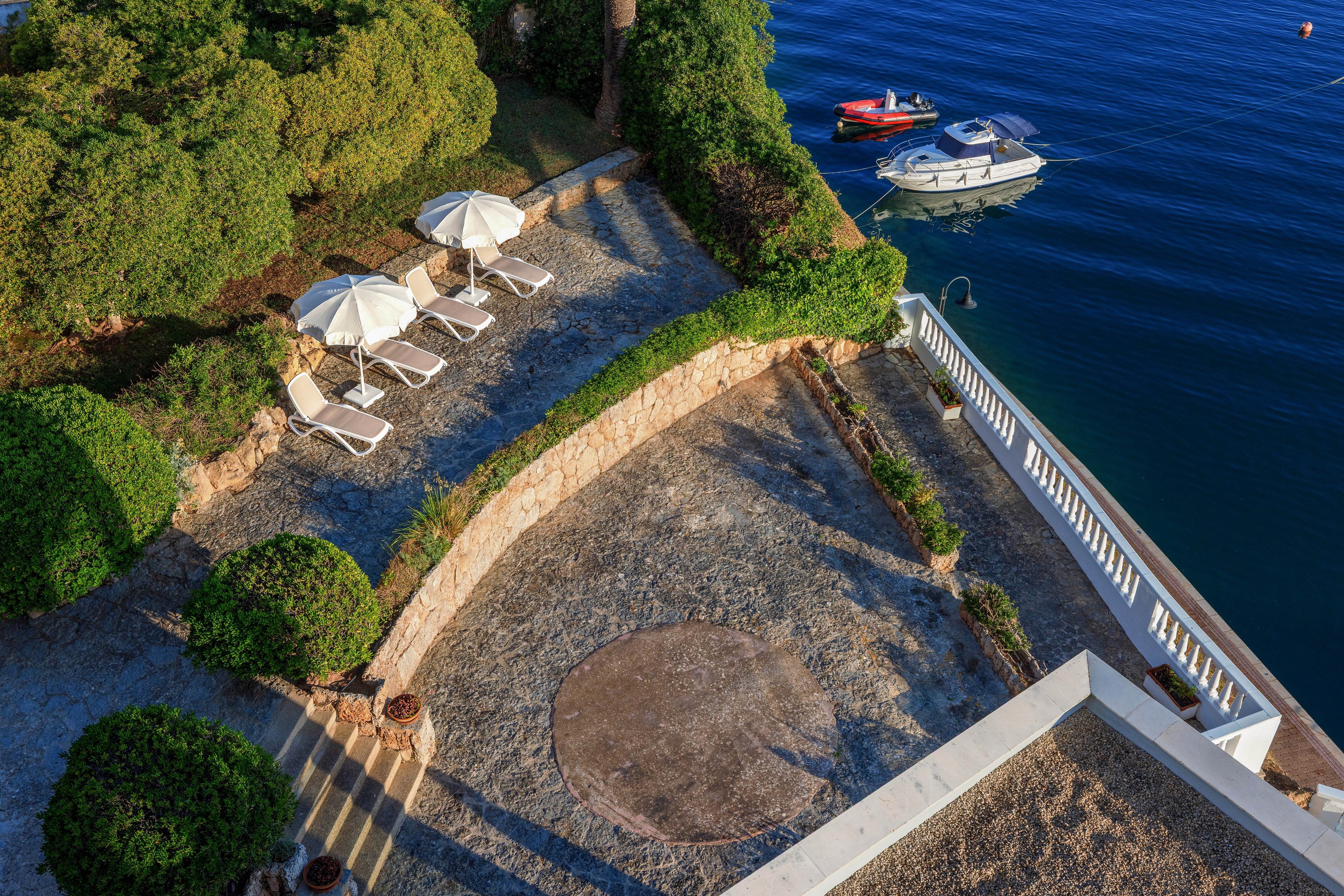 An elevated view shows a stone terrace overlooking calm blue water. Several white sun loungers and umbrellas are arranged on a patio surrounded by manicured green bushes. A white railing separates the terrace from the sea below. Two small boats float nearby, adding to the tranquil coastal atmosphere. Warm sunlight creates long shadows and highlights the textures of the stone and foliage.