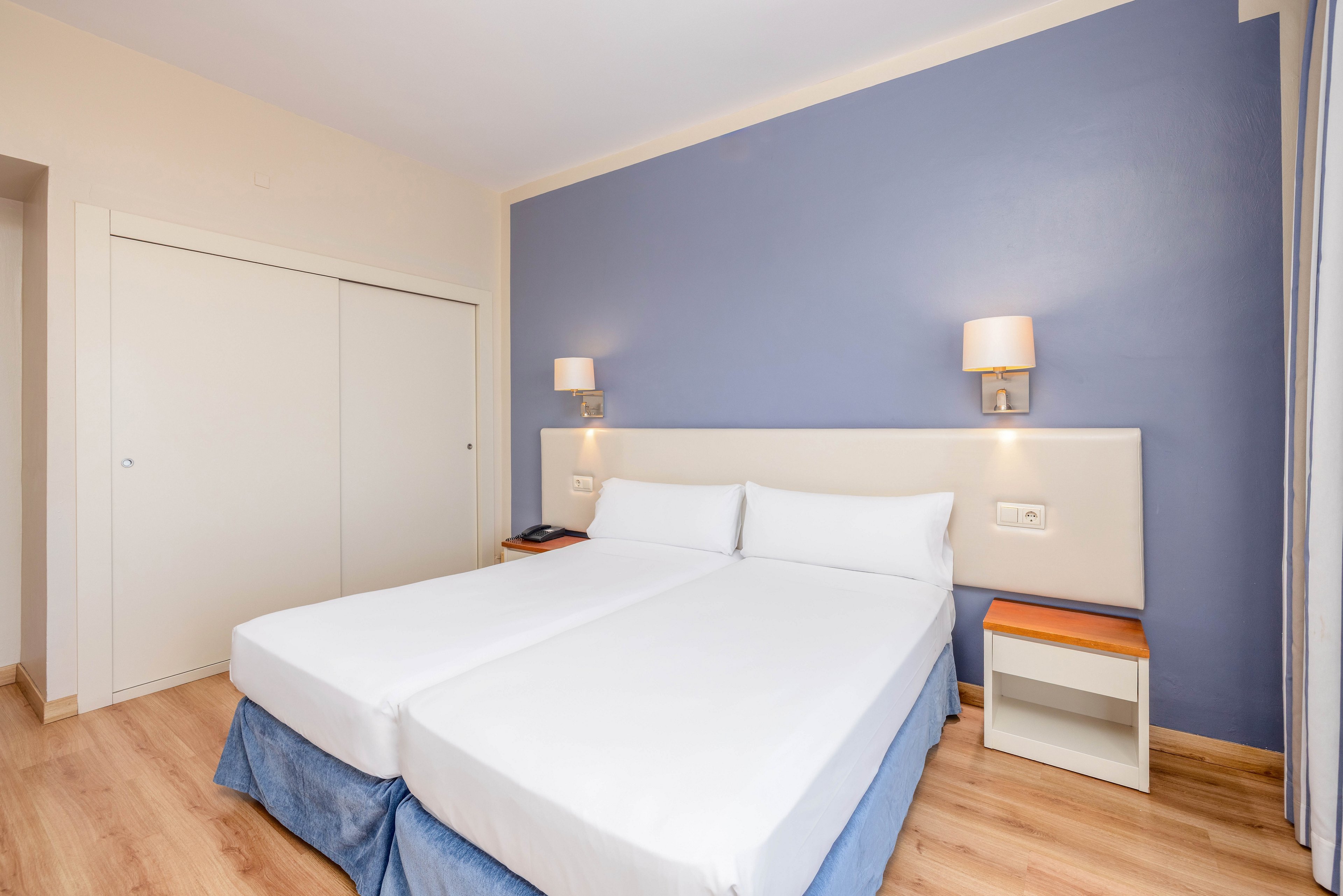 A clean, modern hotel room features two neatly made twin beds pushed together with white linens. The space has a blue accent wall behind the headboard and light wood flooring. Wall-mounted bedside lamps and a small nightstand add functional details. Sliding closet doors and neutral walls contribute to the minimalist, uncluttered atmosphere.