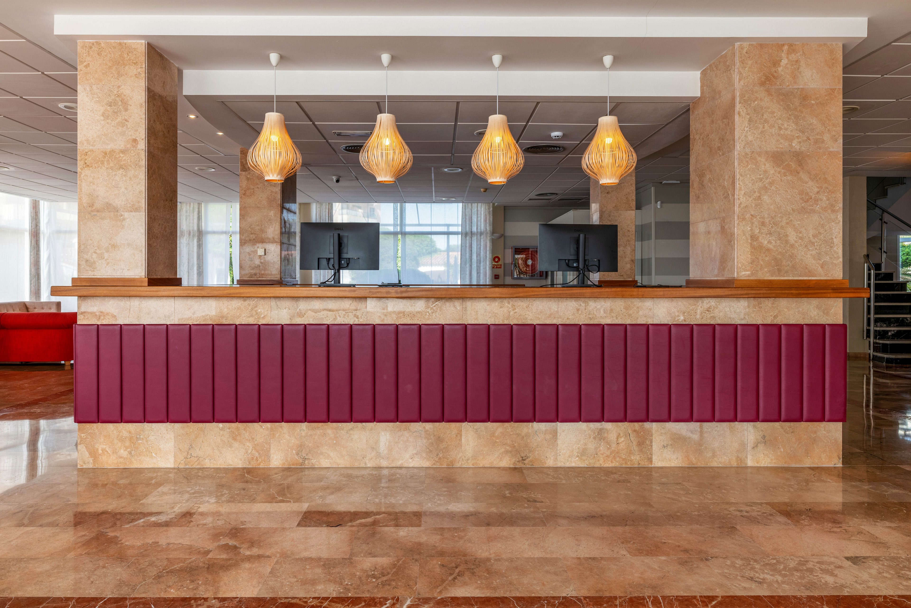 A contemporary hotel reception desk is shown in a spacious lobby. The counter features a burgundy front panel and polished stone surfaces. Four decorative pendant lights hang above the desk, illuminating two computer workstations. Large windows in the background allow natural light to brighten the clean, minimalist interior.