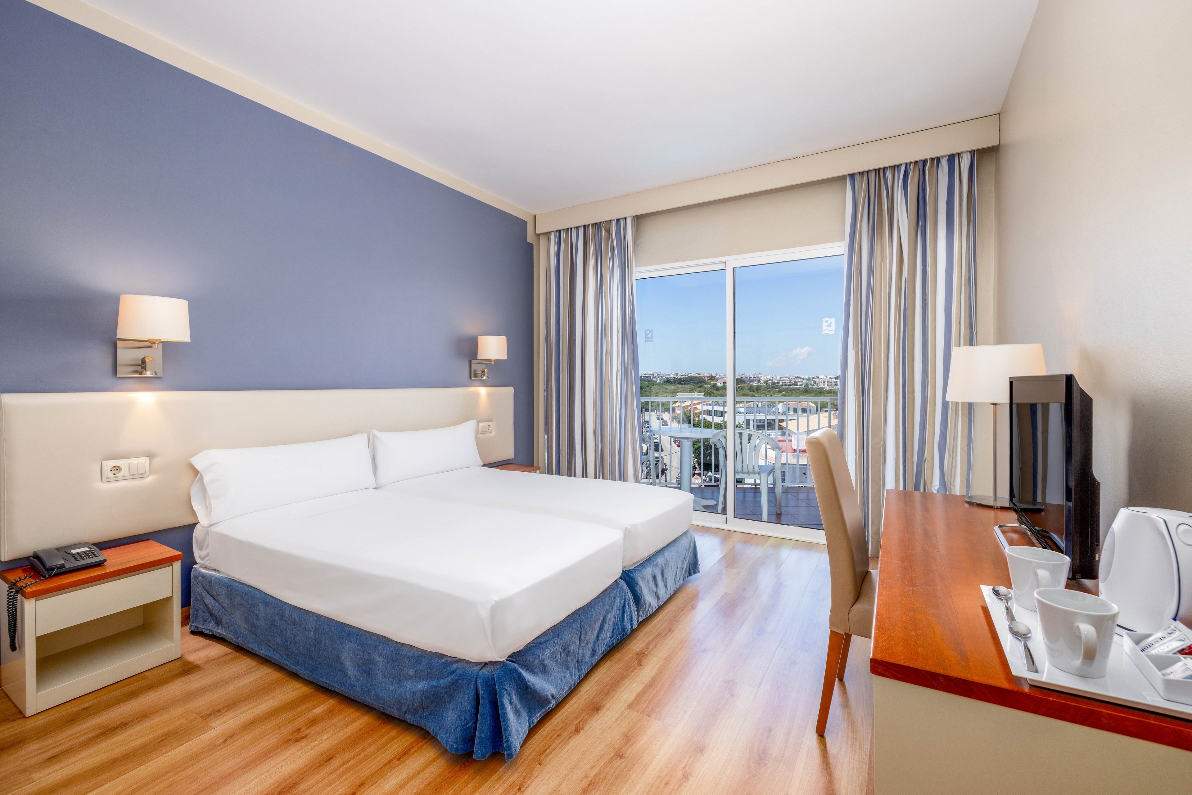 A modern hotel room features a large neatly made bed with white linens and a blue bed skirt. The space includes a wooden desk with a flat-screen TV, telephone, and a tea and coffee service with cups and a kettle. Light wood flooring and blue accent walls create a clean, contemporary look. Large sliding glass doors open to a balcony with a cityscape view under a clear sky, allowing natural light to fill the room.