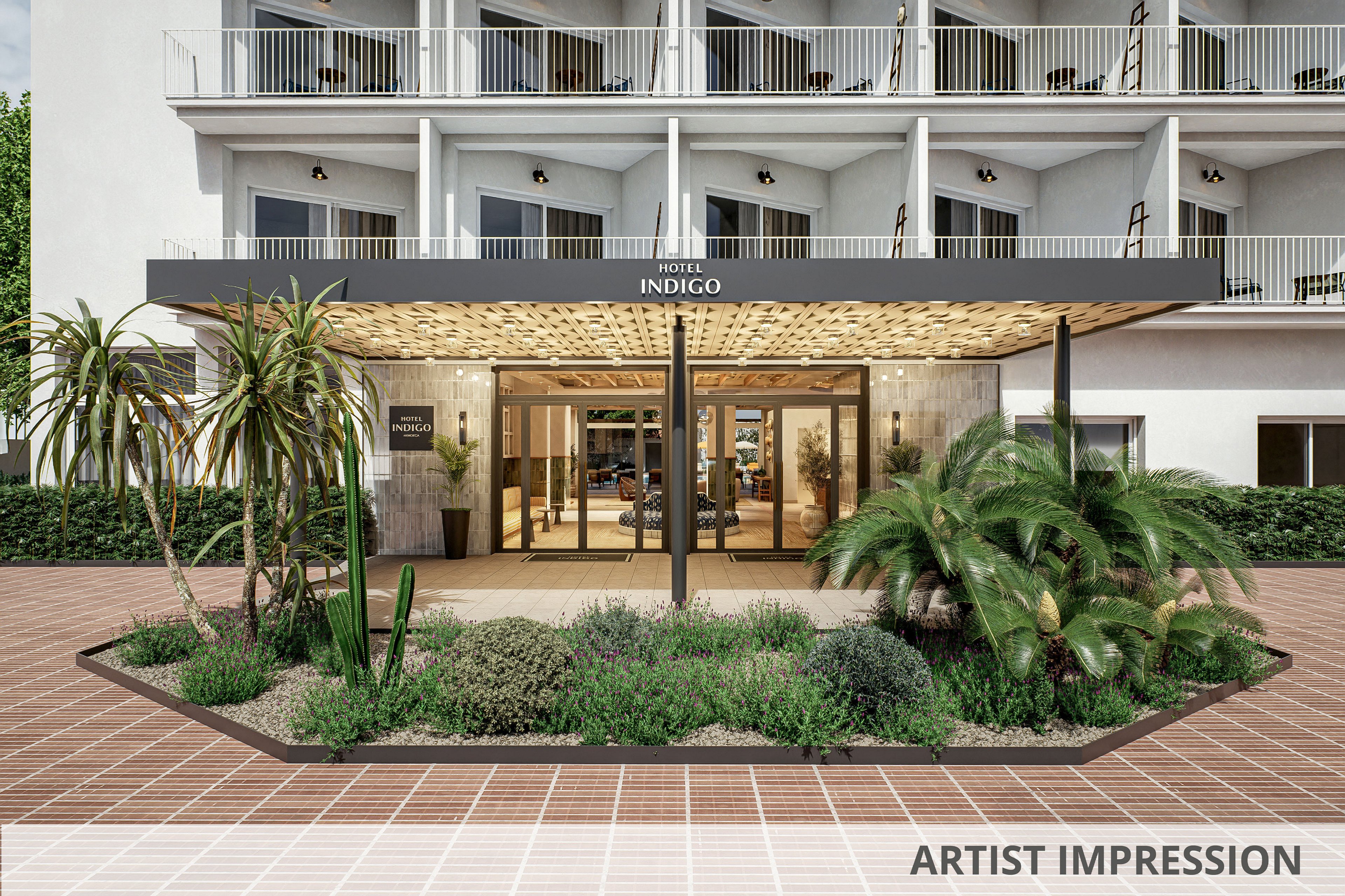 The image shows the front entrance of Hotel Indigo with a modern, welcoming facade. A landscaped garden bed with palm trees, succulents, and low shrubs sits in the foreground on a terracotta-tiled plaza. The hotel’s glass doors reveal a bright lobby area inside, framed by potted plants and warm lighting. Balconies with railings line the upper floors, adding architectural rhythm to the exterior.