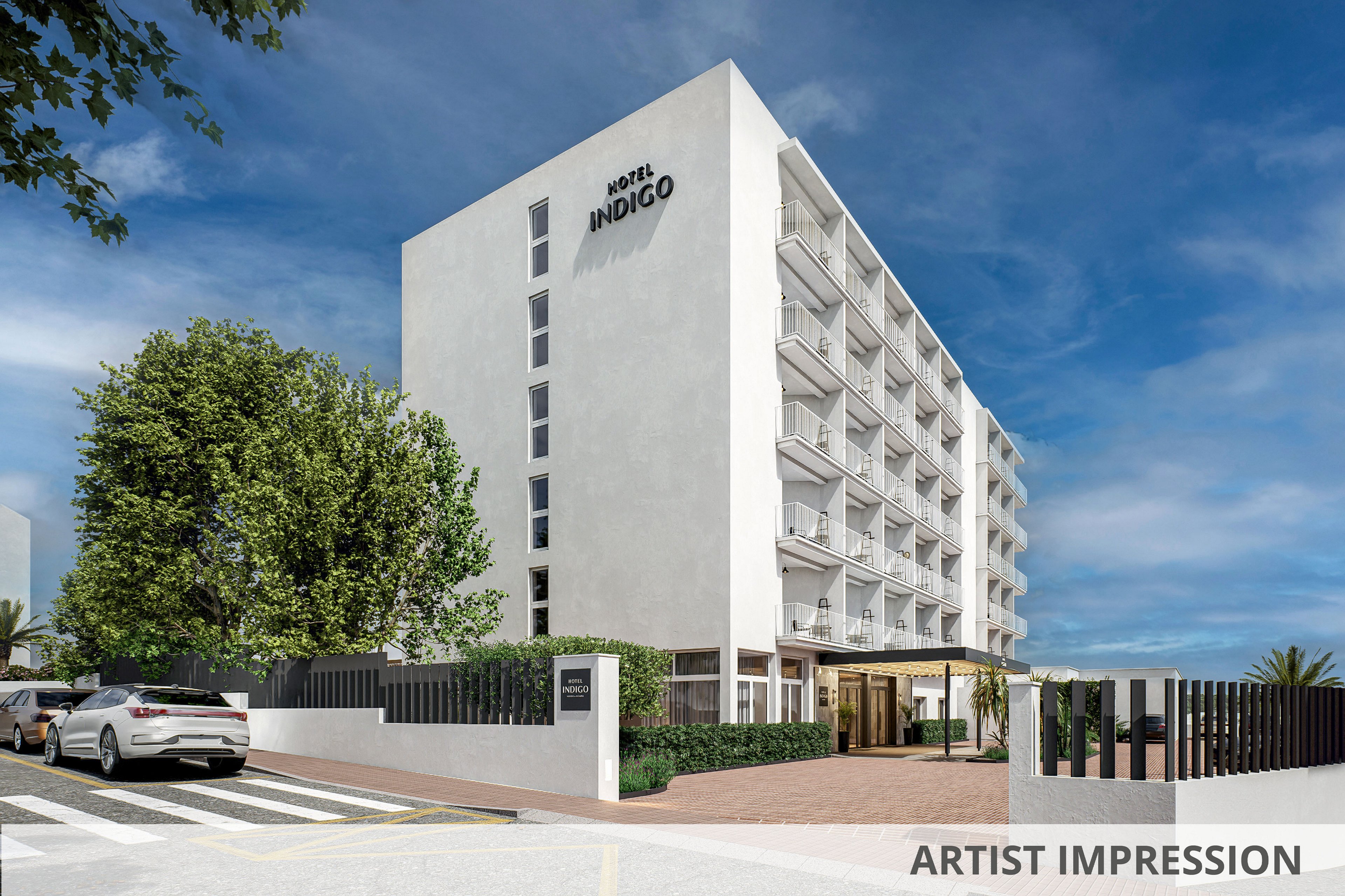 A contemporary multi-story Hotel Indigo building stands on a sunny street corner. The white facade features rows of balconies and large windows, with the hotel entrance visible at ground level. Several parked cars line the road, and a crosswalk and sidewalk frame the scene. Lush green trees and a clear blue sky add a bright, welcoming atmosphere.