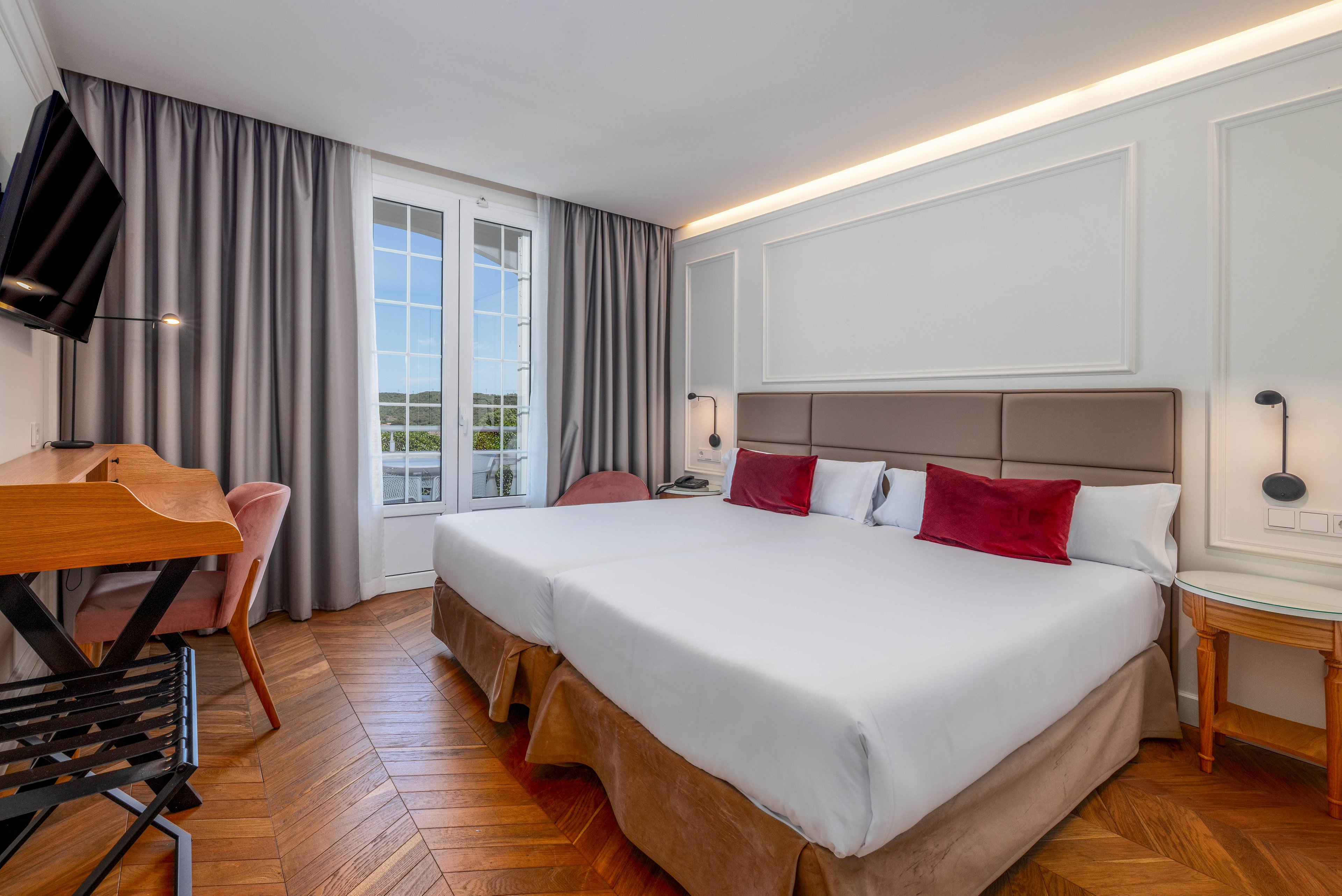 A contemporary hotel room features two neatly made twin beds with white linens and red accent pillows. The space includes a wooden desk with a chair, a wall-mounted television, and a small bedside table with a lamp. Large French doors with grid windows open to an outdoor view, framed by long gray curtains. Warm wood flooring and soft neutral tones create a clean, inviting atmosphere.