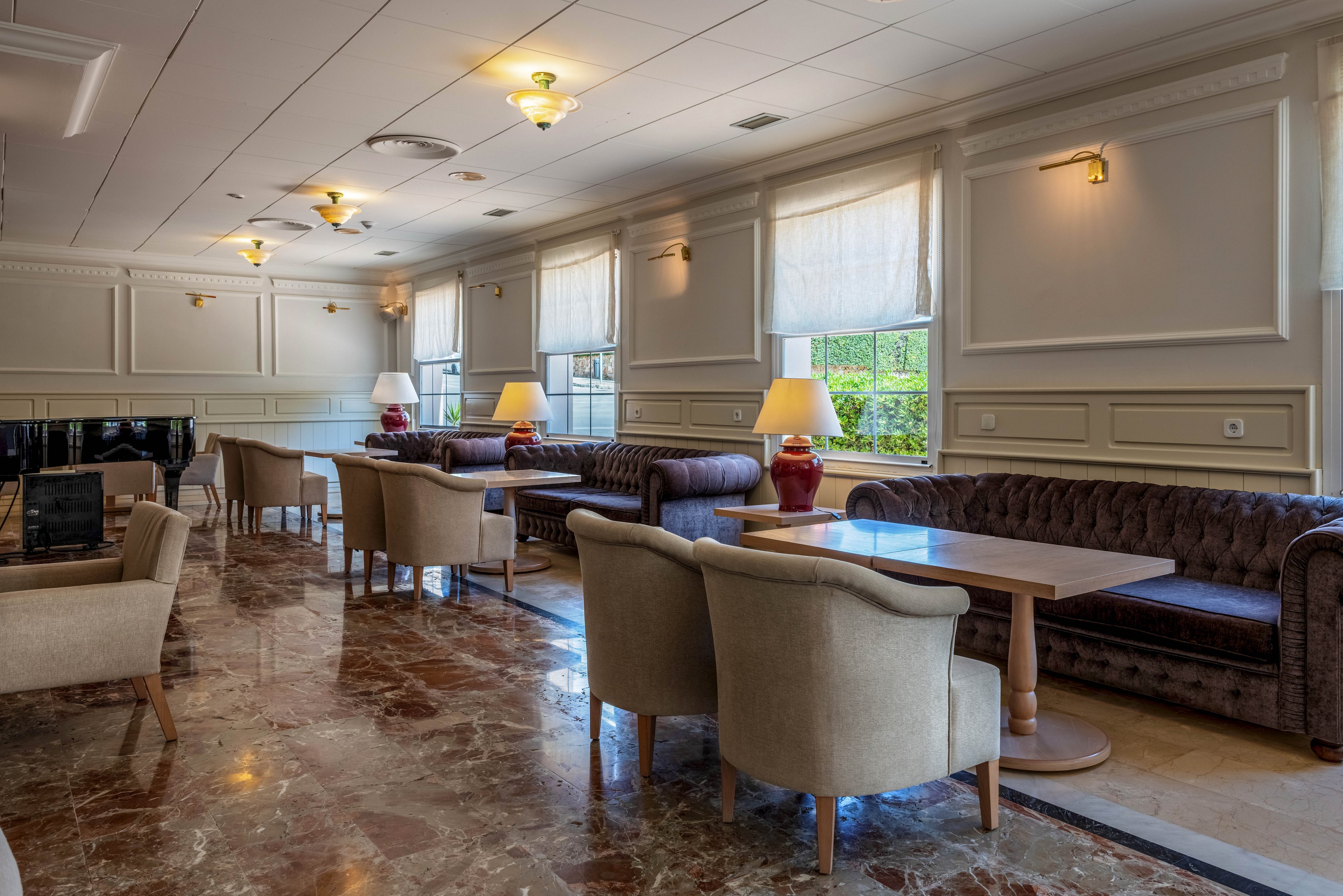 A sophisticated lounge area features plush armchairs and tufted leather sofas arranged around wooden tables. Large windows with sheer blinds allow natural light to brighten the room and reveal greenery outside. Warm wall sconces and table lamps create a cozy, inviting atmosphere despite the space being empty. The polished marble floor and classic wall paneling add to the upscale, refined interior design.