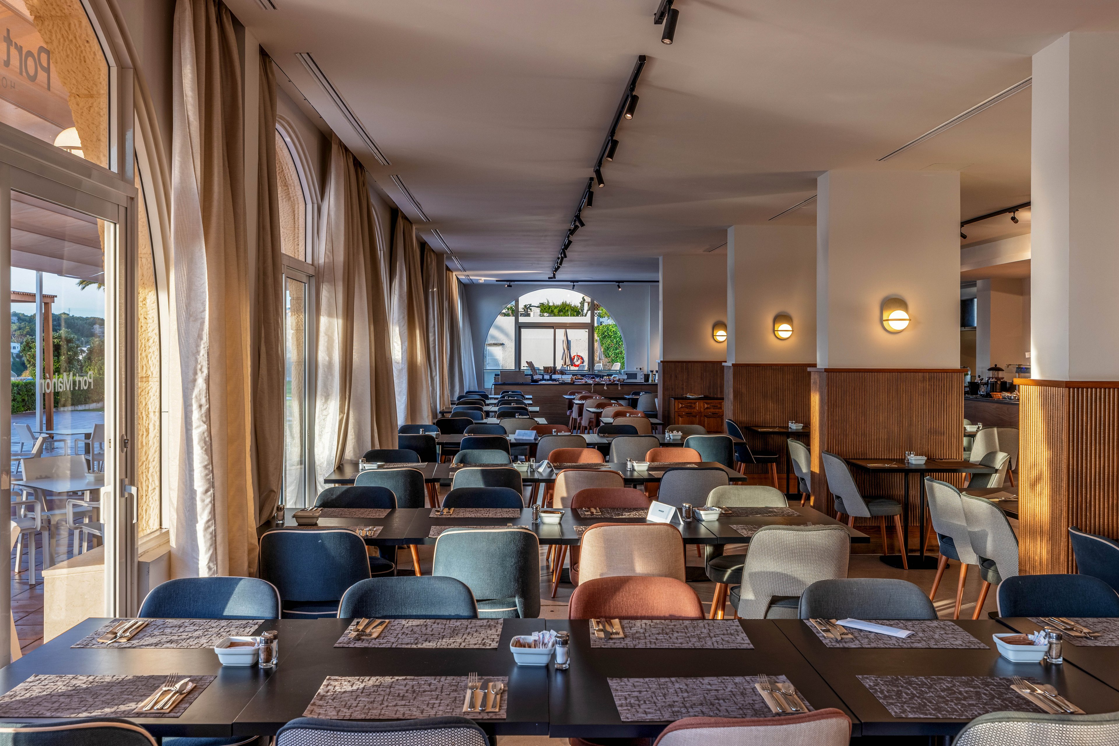 Spacious restaurant dining room with neatly arranged tables and chairs is shown in warm afternoon light. Large arched windows with flowing curtains line one side, opening to an outdoor terrace and distant view. Each table is set with placemats, cutlery, and small condiment containers, ready for guests. Soft wall sconces and ceiling track lights create a cozy yet contemporary atmosphere in the empty space.