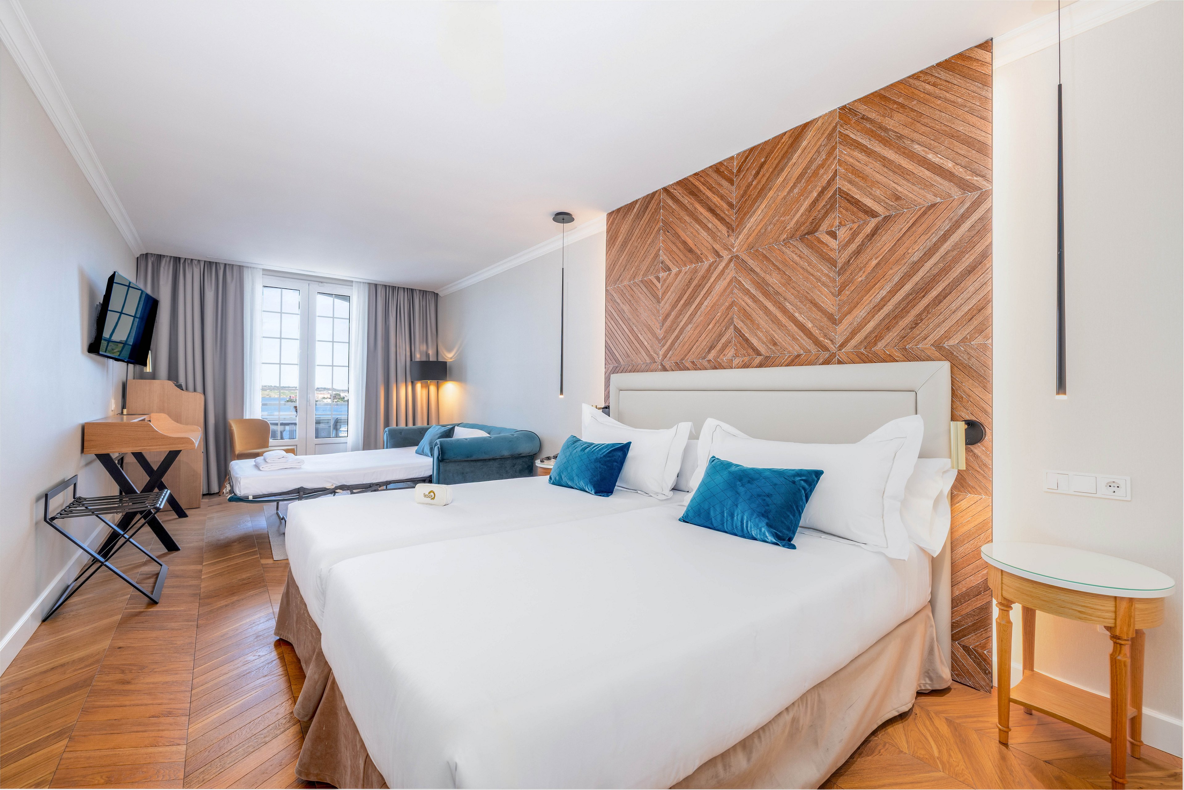 Spacious hotel room featuring two large neatly made beds with white linens and blue accent pillows. The space includes a wooden herringbone accent wall, hardwood flooring, and contemporary pendant lighting. A wall-mounted television, small desk, and additional seating area with a sofa and extra bed are visible. Large windows with curtains allow natural light to brighten the clean, minimalist interior.
