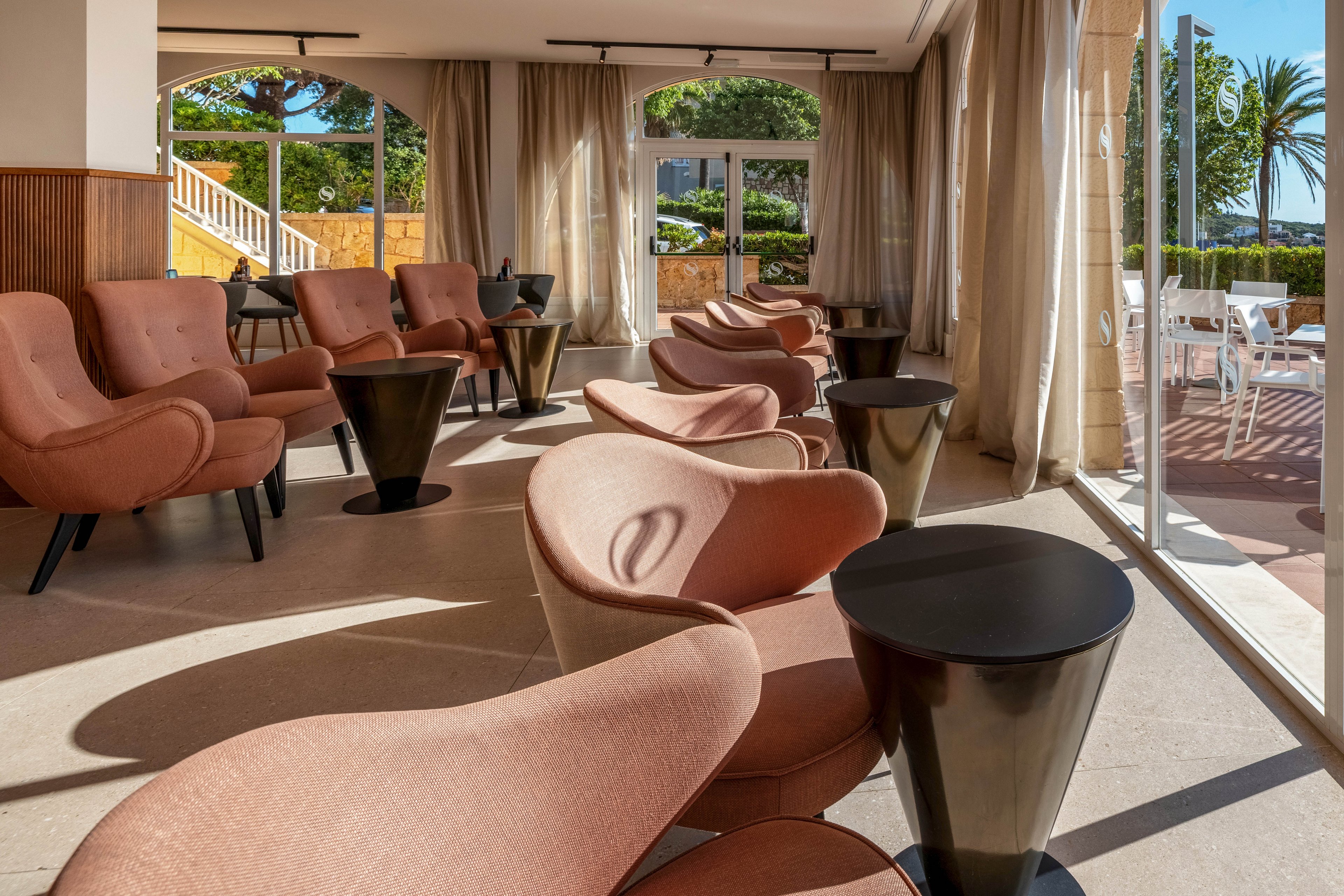 A bright hotel lounge features rows of contemporary pink armchairs paired with small black side tables. Large floor-to-ceiling windows let in abundant sunlight and offer views of an outdoor terrace with greenery and palm trees. Sheer curtains frame the windows, softening the light and creating a relaxed atmosphere. The space appears quiet and empty, ideal for casual meetings or peaceful relaxation.
