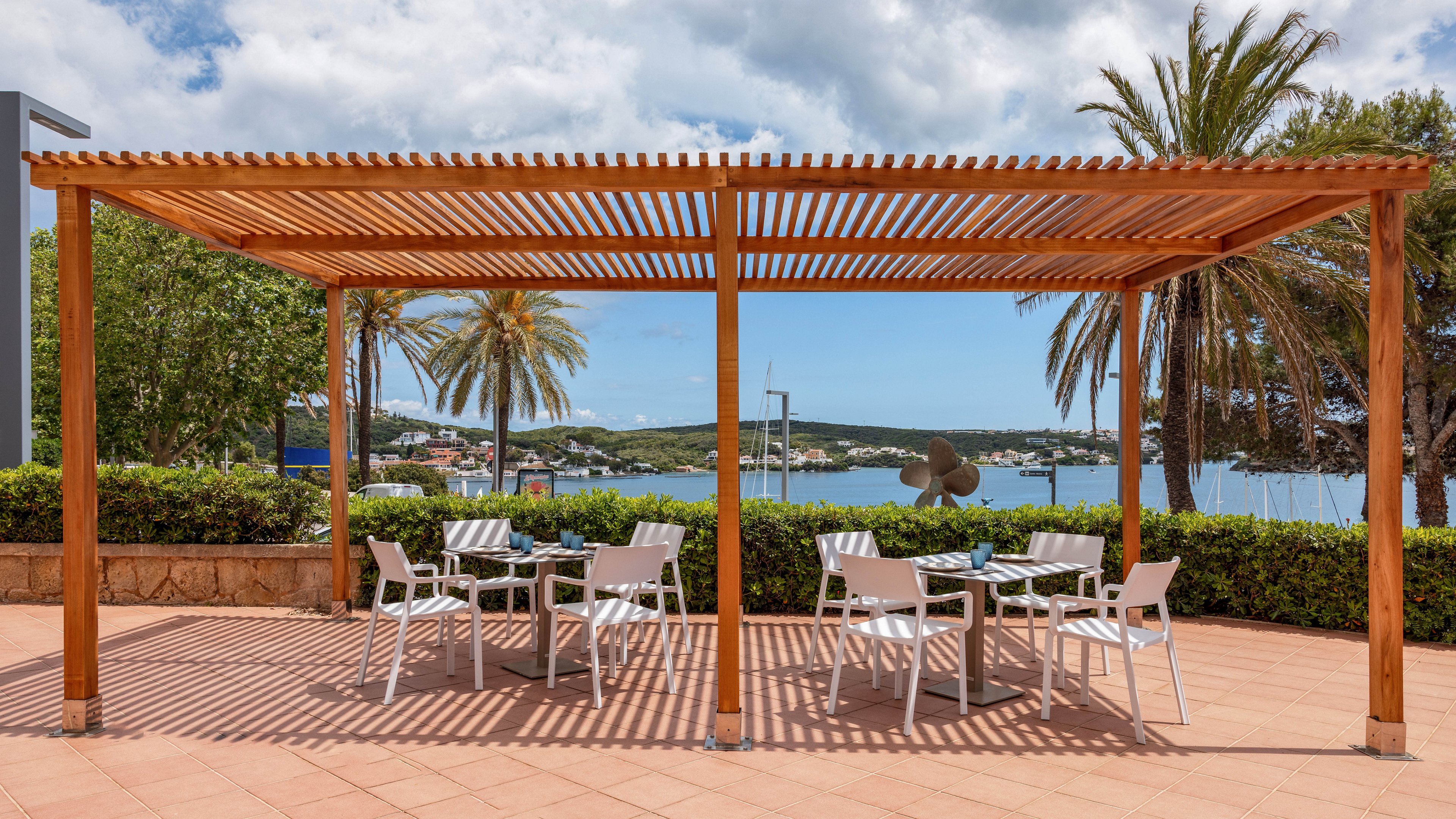A wooden pergola shades several white tables and chairs on a tiled outdoor terrace. The seating area overlooks a calm waterfront with boats and low buildings in the distance. Lush green hedges and tall palm trees frame the scene under a partly cloudy sky. The setting suggests a relaxed coastal café or lounge area ideal for outdoor dining or leisure.