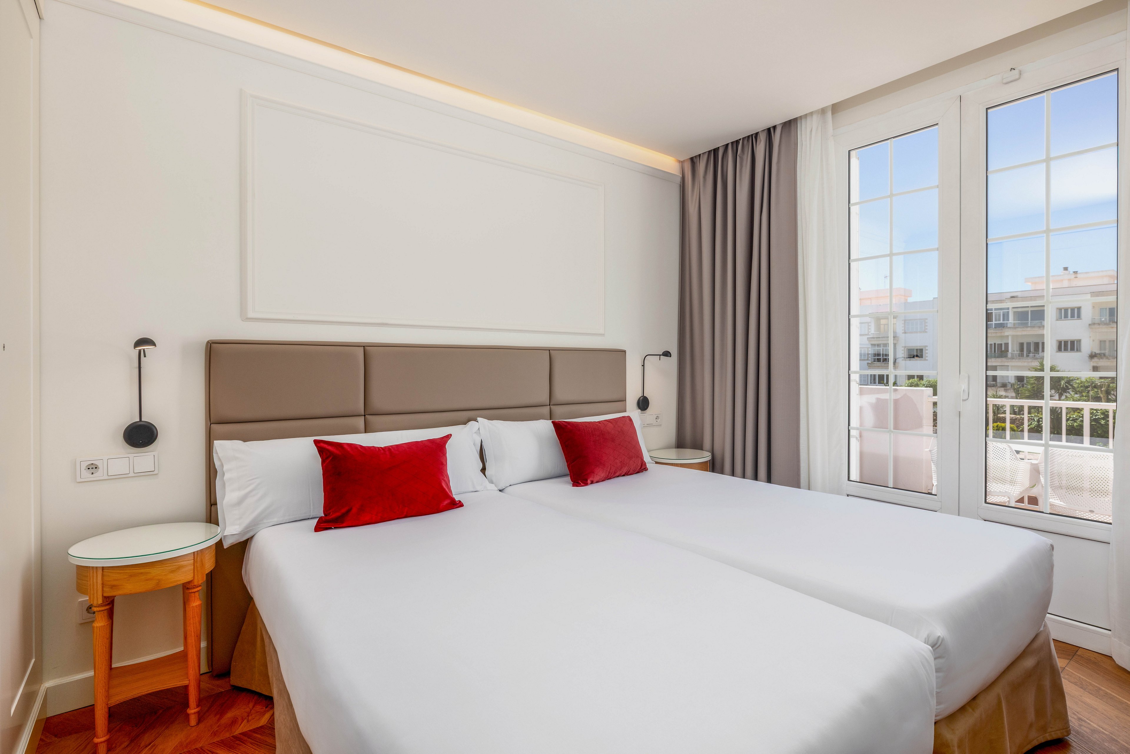 A modern hotel room features two neatly made twin beds with white linens and red accent pillows. The space is bright, with natural light streaming in through large glass doors that open to a balcony. Minimalist decor includes a padded headboard, bedside tables, and wall-mounted reading lights. Neutral walls and curtains create a calm, contemporary atmosphere suitable for travelers or vacation stays.