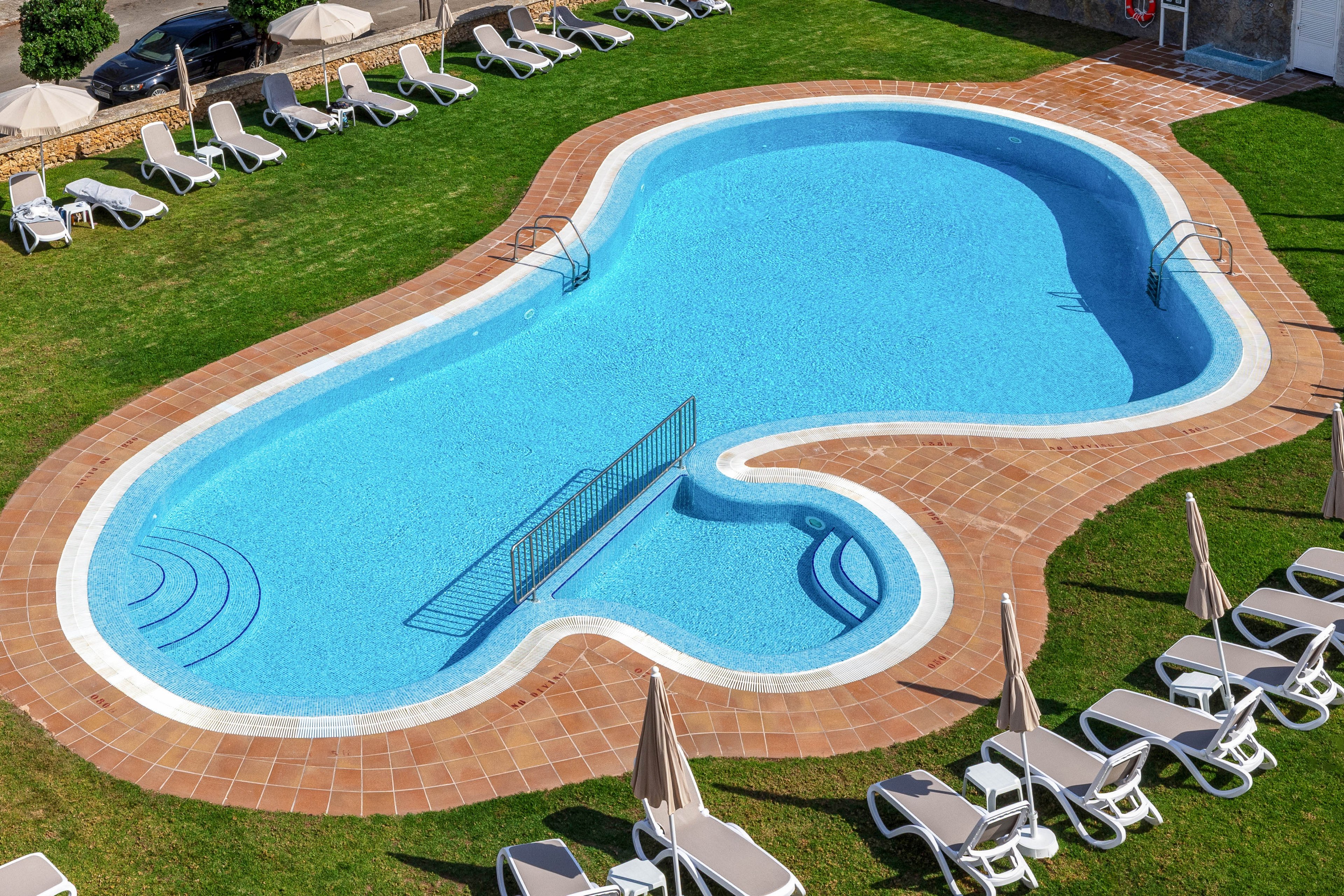 A uniquely shaped outdoor swimming pool is surrounded by a tiled deck and green lawn. White sun loungers and closed beige umbrellas are neatly arranged around the pool, suggesting a resort or hotel setting. The clear blue water reveals built-in steps and a metal handrail for easy access. No people are present, creating a calm and orderly atmosphere ideal for relaxation.