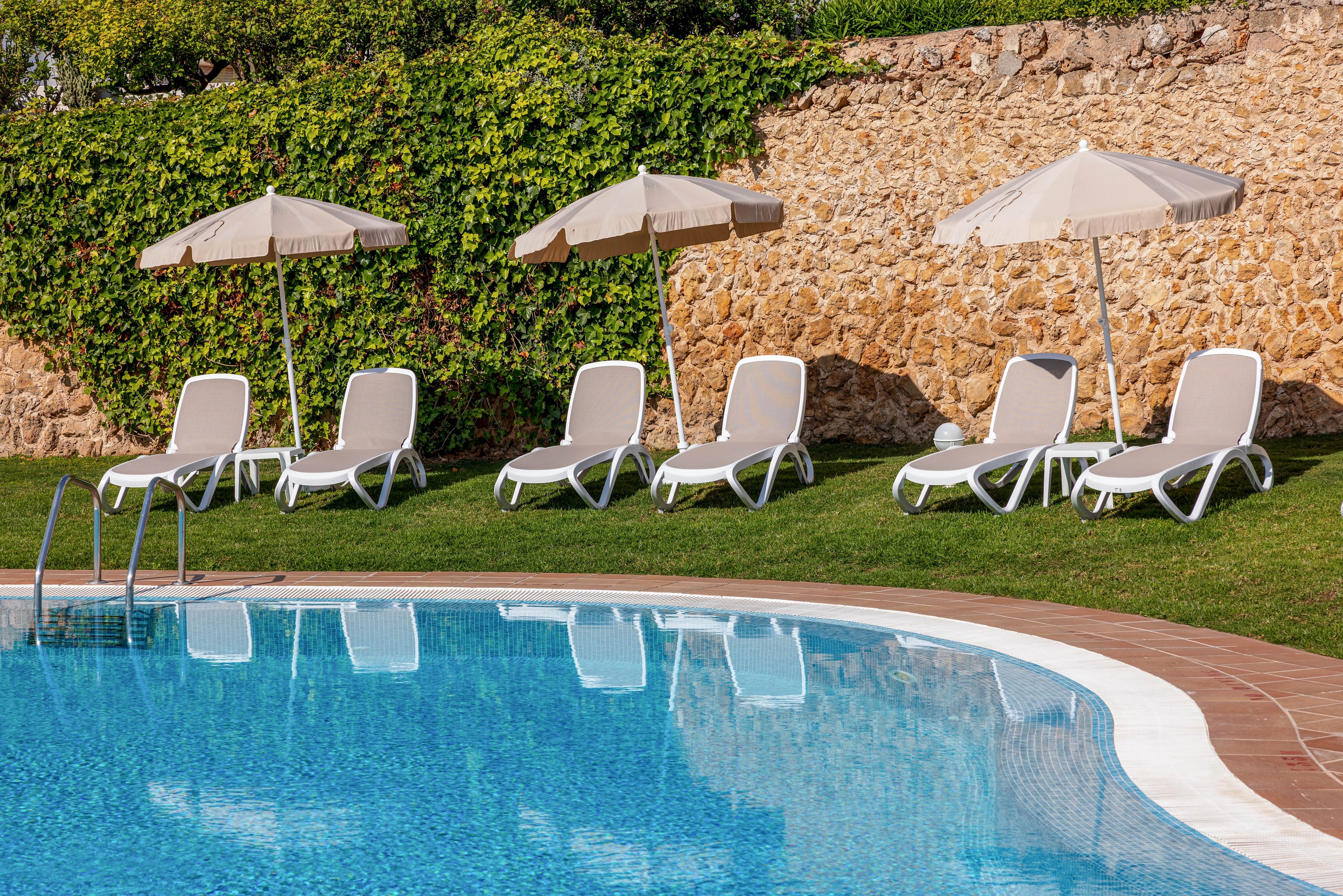 A clear blue swimming pool sits in the foreground, reflecting the sunlight. Along the grassy edge, several white sun loungers are neatly arranged beneath matching parasols. A stone wall covered partly with green ivy forms the backdrop, creating a private and tranquil atmosphere. The scene suggests a quiet resort or hotel pool area ready for guests.