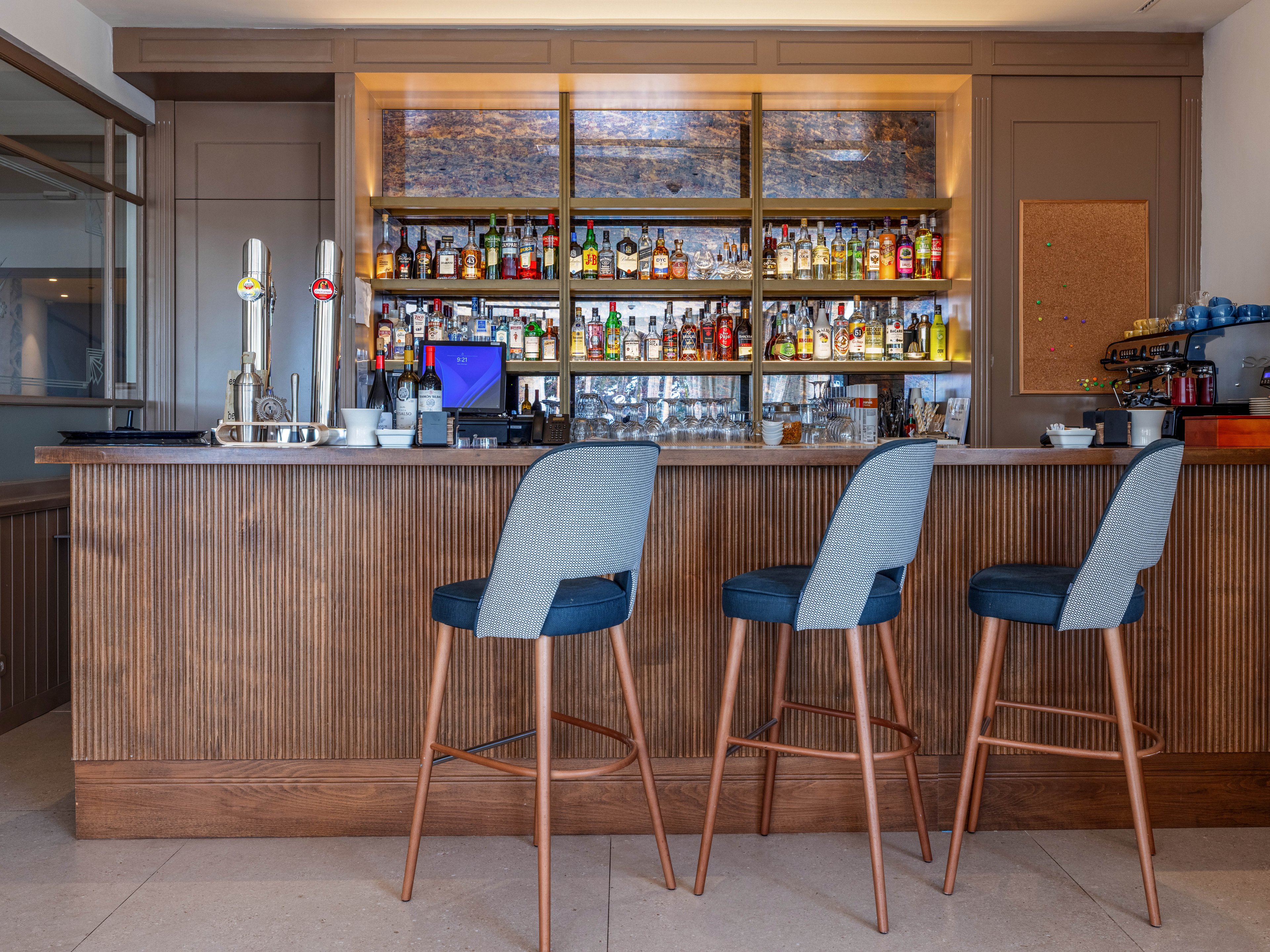 A contemporary bar counter is shown with three upholstered bar stools neatly arranged in front. The back wall features shelves filled with assorted liquor bottles and glassware, creating a colorful focal point. Beer taps, cups, and a small coffee station are visible on the countertop, suggesting a versatile drink service area. The overall setting appears clean and inviting, with warm wood finishes and neutral tones contributing to a relaxed atmosphere.