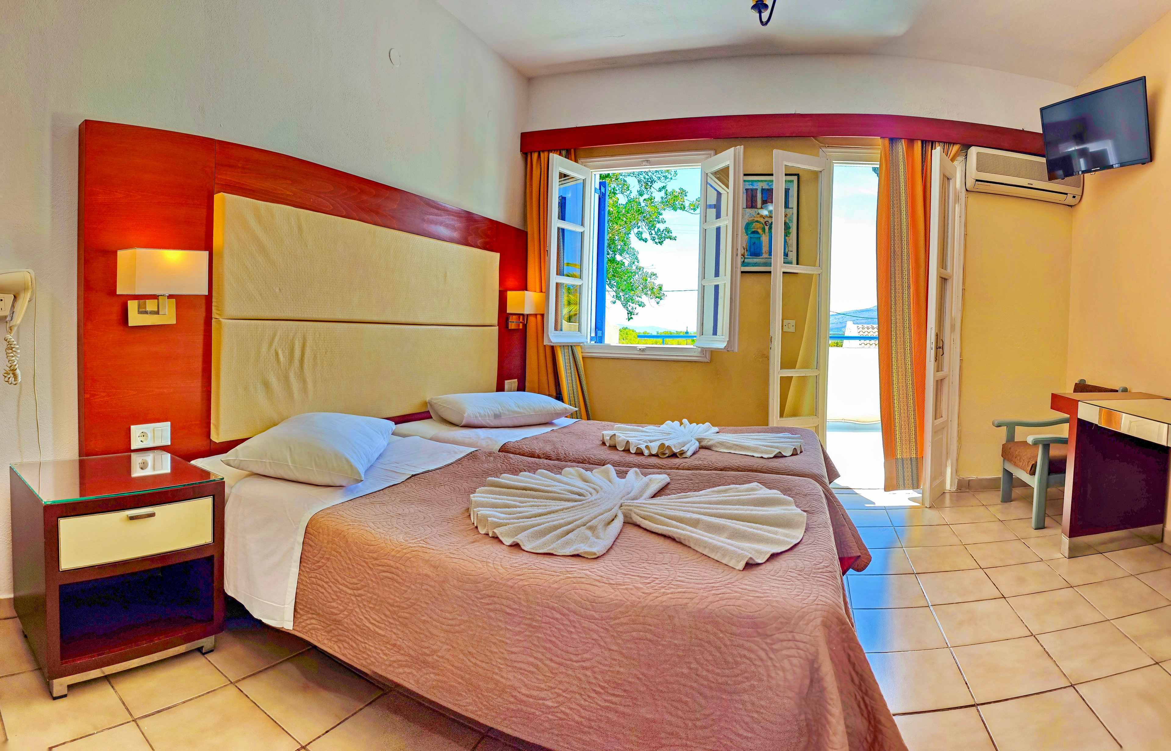 A tidy hotel room features two twin beds pushed together, topped with folded towel decorations. The space is bright and airy, with tiled floors and light-colored walls. An open balcony door and windows reveal greenery and a glimpse of the outdoors, allowing natural light to fill the room. A wall-mounted television, air conditioner, bedside tables, and a small desk complete the comfortable interior.