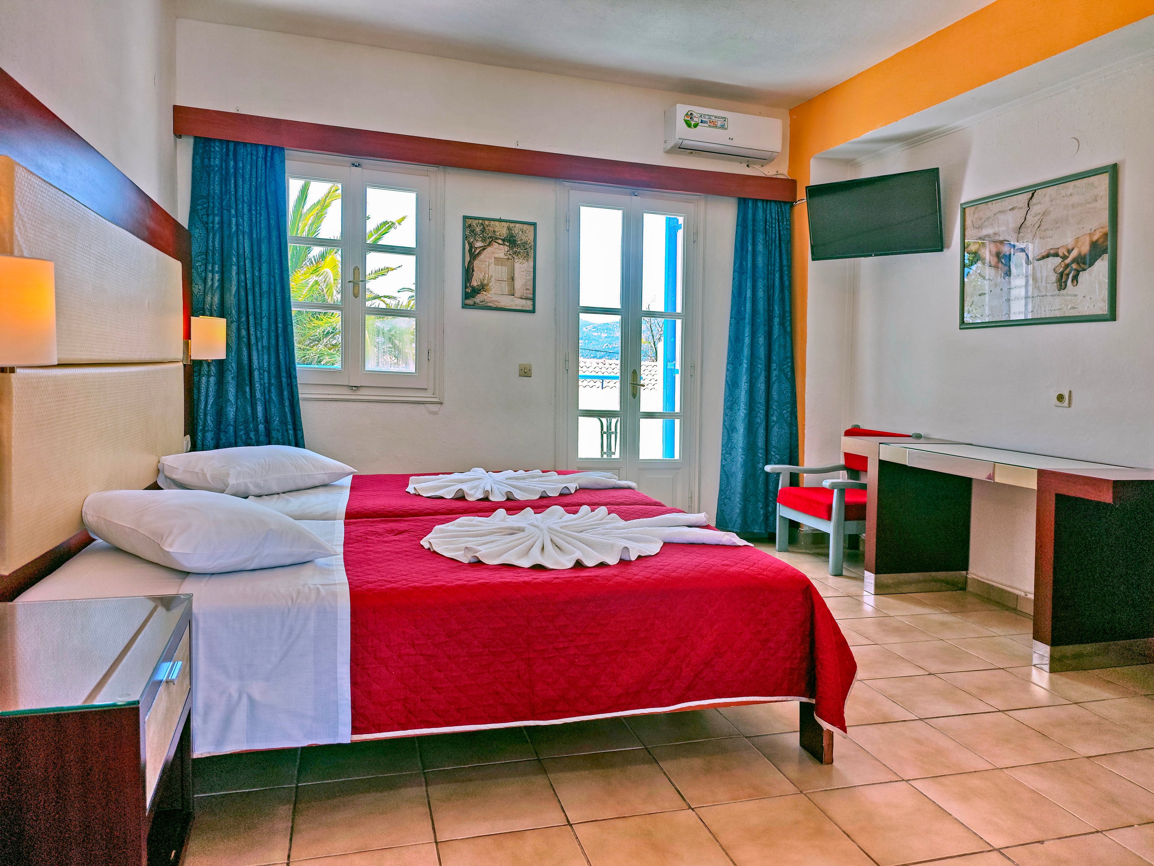 A colorful hotel room features two neatly made twin beds with red bedspreads and folded white towels arranged decoratively on top. Large windows and a glass door with blue curtains let in natural light and offer a view outside. The room includes a wall-mounted television, a desk with a chair, and framed artwork on the walls. Tiled flooring and simple modern furniture create a clean, inviting atmosphere suitable for vacation or business travel.