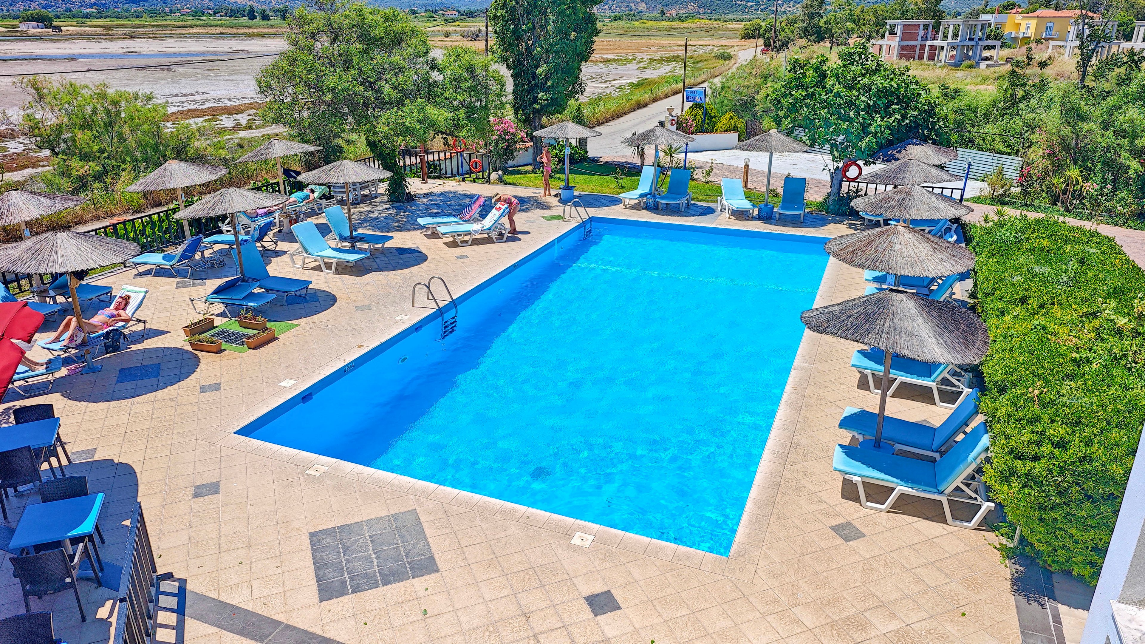 An outdoor swimming pool is surrounded by sun loungers and straw parasols at a holiday resort. The bright blue water contrasts with the beige tiled pool deck and scattered seating areas. A few people relax near the pool while greenery and distant buildings frame the background. The scene suggests a warm, sunny day ideal for vacation and leisure.