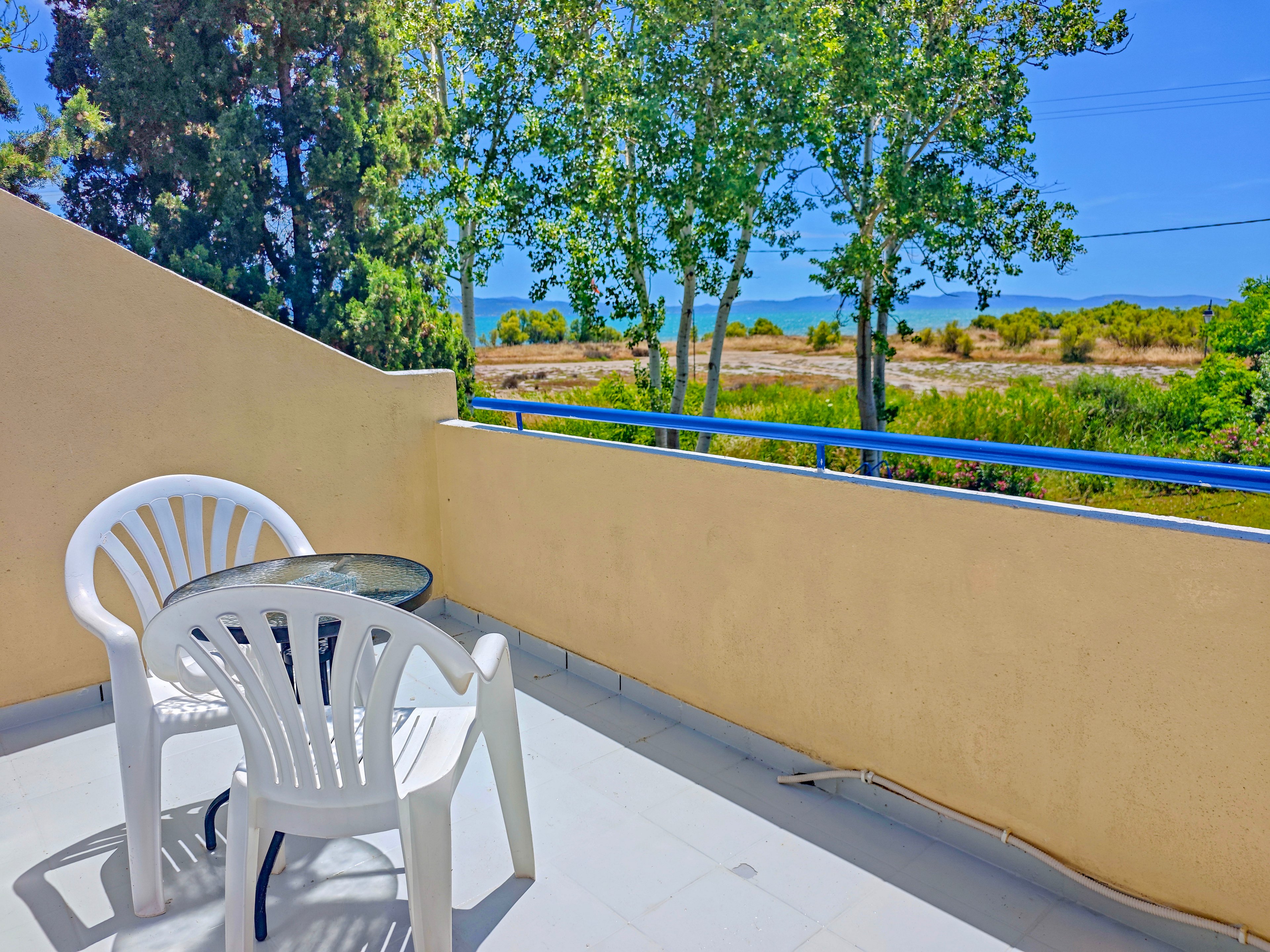 A small balcony features a round glass-top table and two white plastic chairs. The space overlooks a natural landscape with trees, shrubs, and a glimpse of the sea in the distance. Bright sunlight casts sharp shadows on the clean tiled floor, emphasizing the warm, clear weather. The scene conveys a peaceful outdoor setting ideal for relaxing or enjoying a quiet drink.