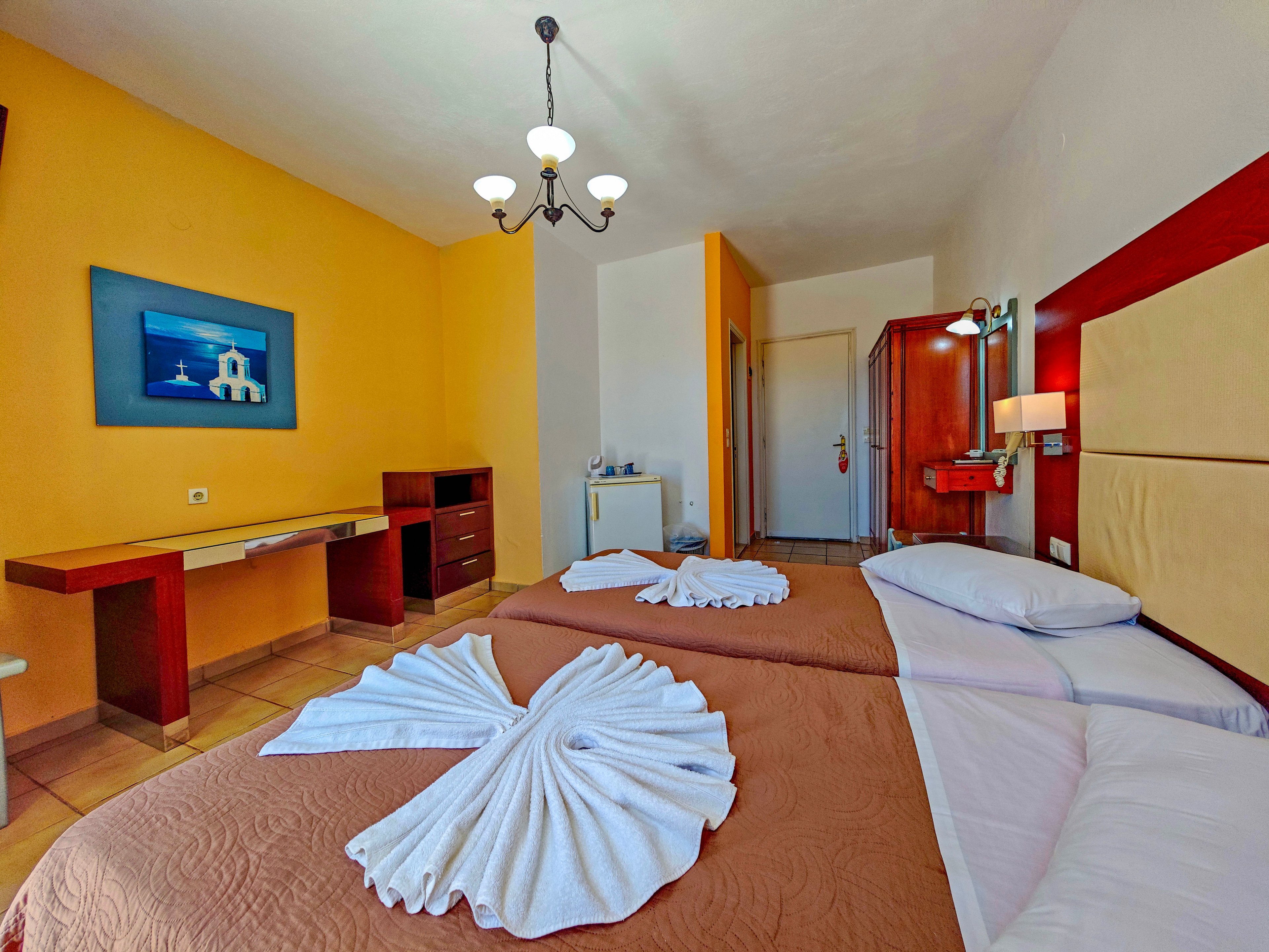 A cozy hotel room features two neatly made twin beds with brown covers and white towels folded into decorative shapes. The space is brightly lit, with a warm yellow accent wall contrasting against neutral tones. A desk, chair, and small refrigerator are visible along one side, while a wall-mounted lamp and framed artwork add character. The overall atmosphere is clean, inviting, and suitable for vacation or business travel.