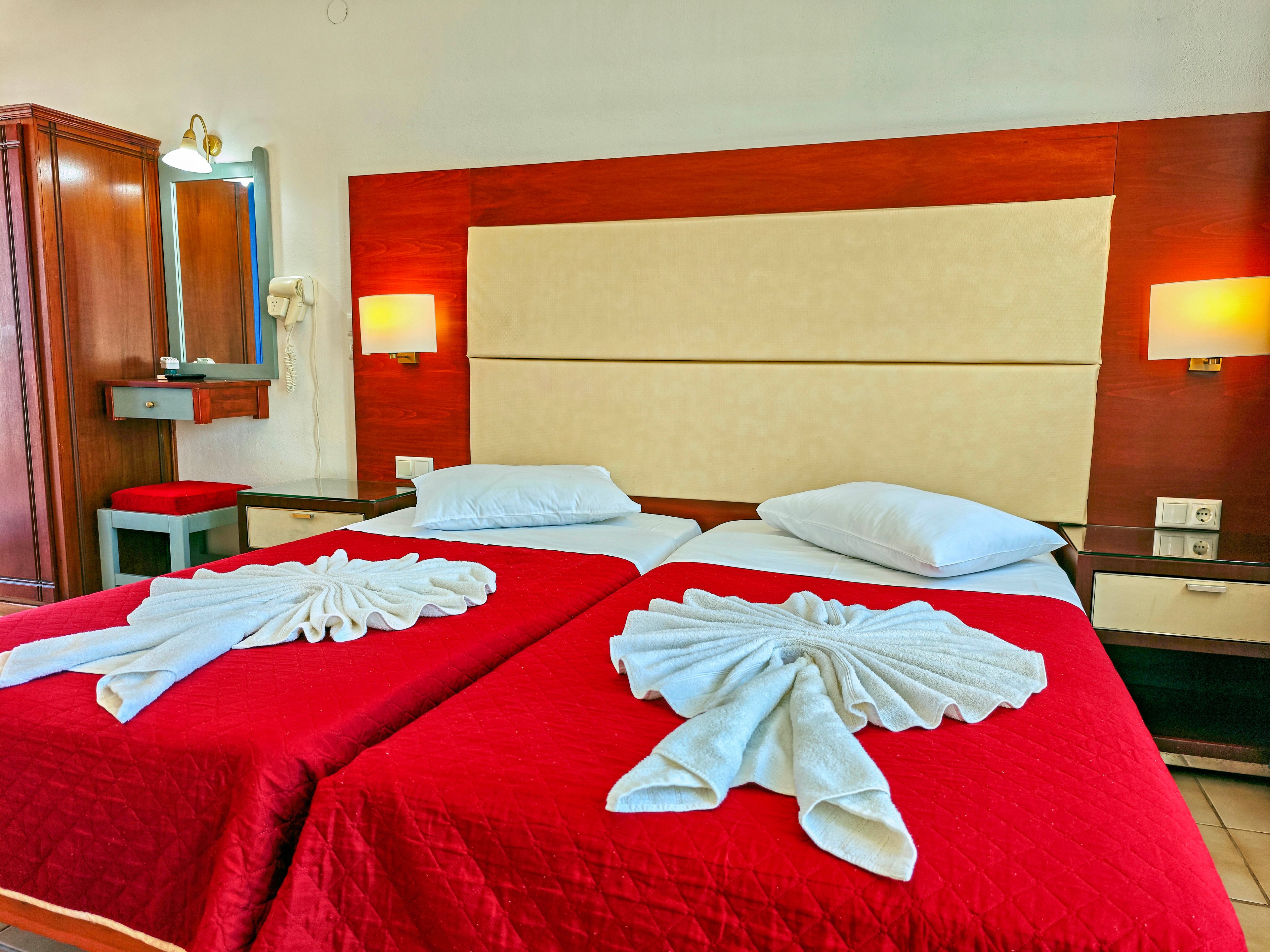 A hotel room features two neatly made twin beds with bright red bedspreads and white pillows. Decorative white towels folded into swan shapes rest at the foot of each bed, adding a welcoming touch. The headboard is upholstered in a light beige panel against a wooden accent wall. Warm wall-mounted lamps, a dressing table with mirror, and a small stool complete the cozy interior setting.