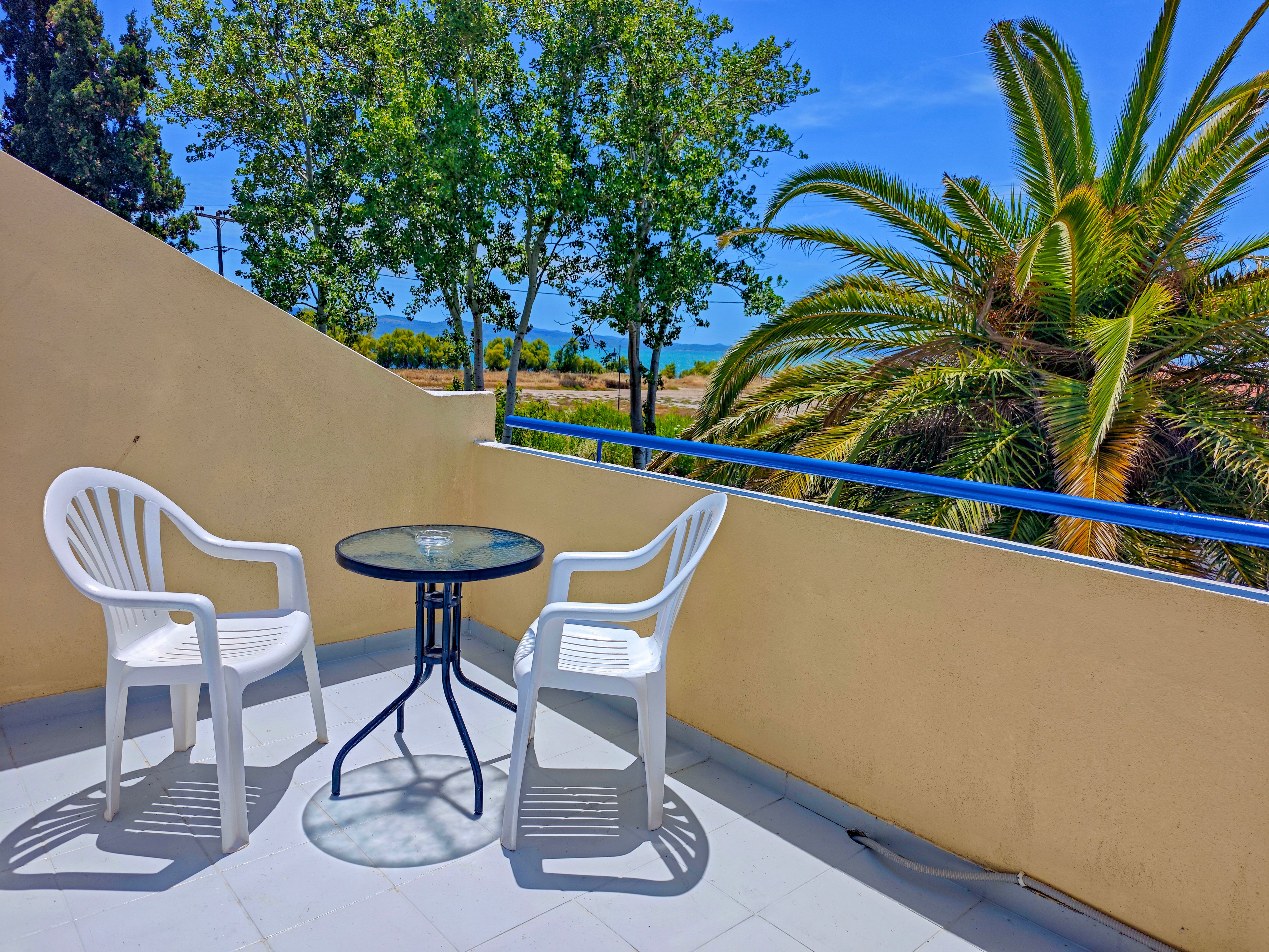 An outdoor balcony features two white plastic chairs around a small round glass-top table. The space is enclosed by a light-colored wall and overlooks lush green trees and a tall palm tree. Bright sunlight casts sharp shadows on the tiled floor, suggesting a warm, clear day. In the distance, a glimpse of blue sky and landscape adds to the relaxed, vacation-like atmosphere.