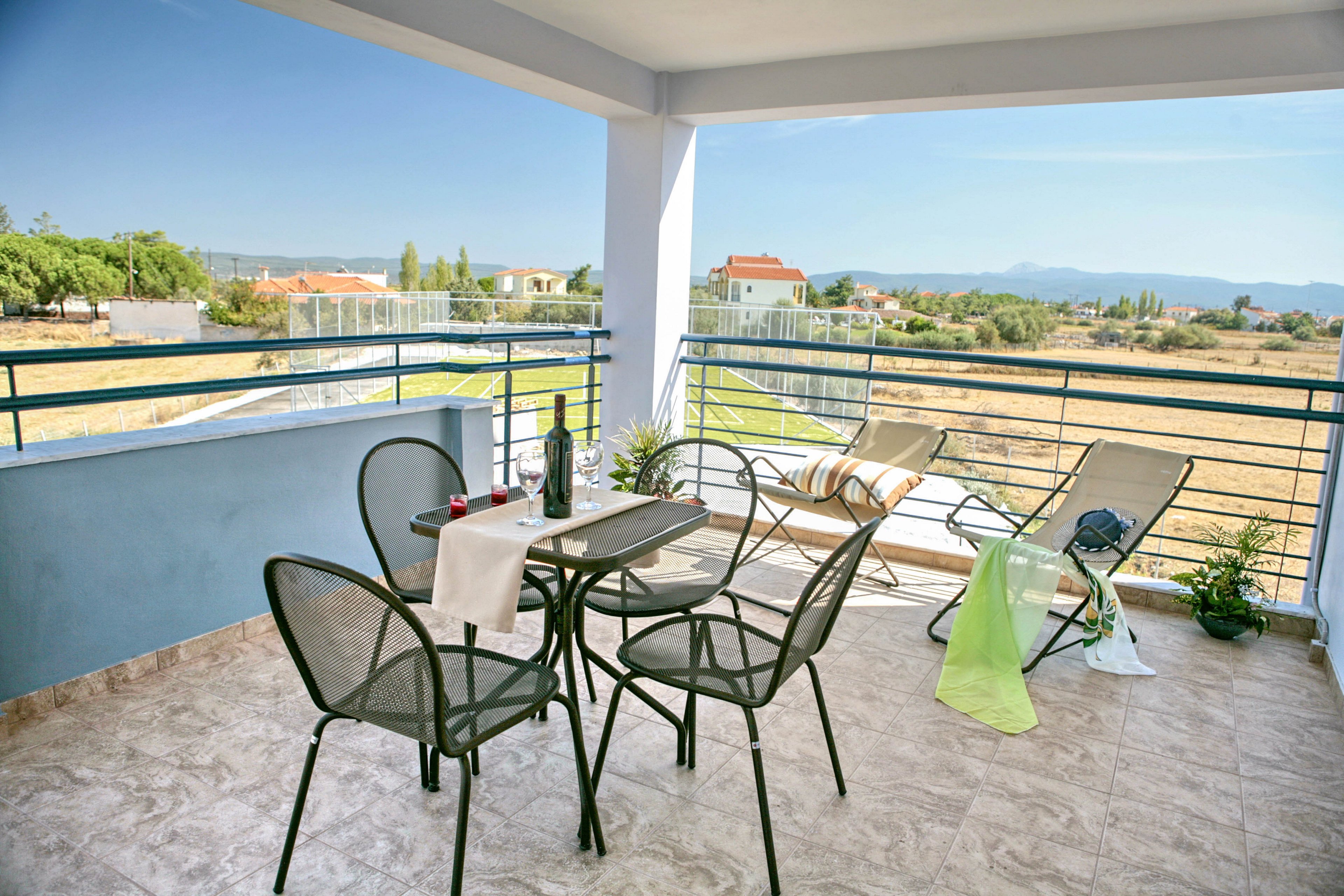 An open terrace features a small table with four mesh chairs and a bottle with glasses arranged on top. Two reclining lounge chairs with towels are positioned near the railing, facing a wide rural landscape. The scene is bright and airy, with tiled flooring, potted plants, and distant houses under a clear blue sky. The overall atmosphere suggests a relaxing outdoor space for leisure and scenic views.