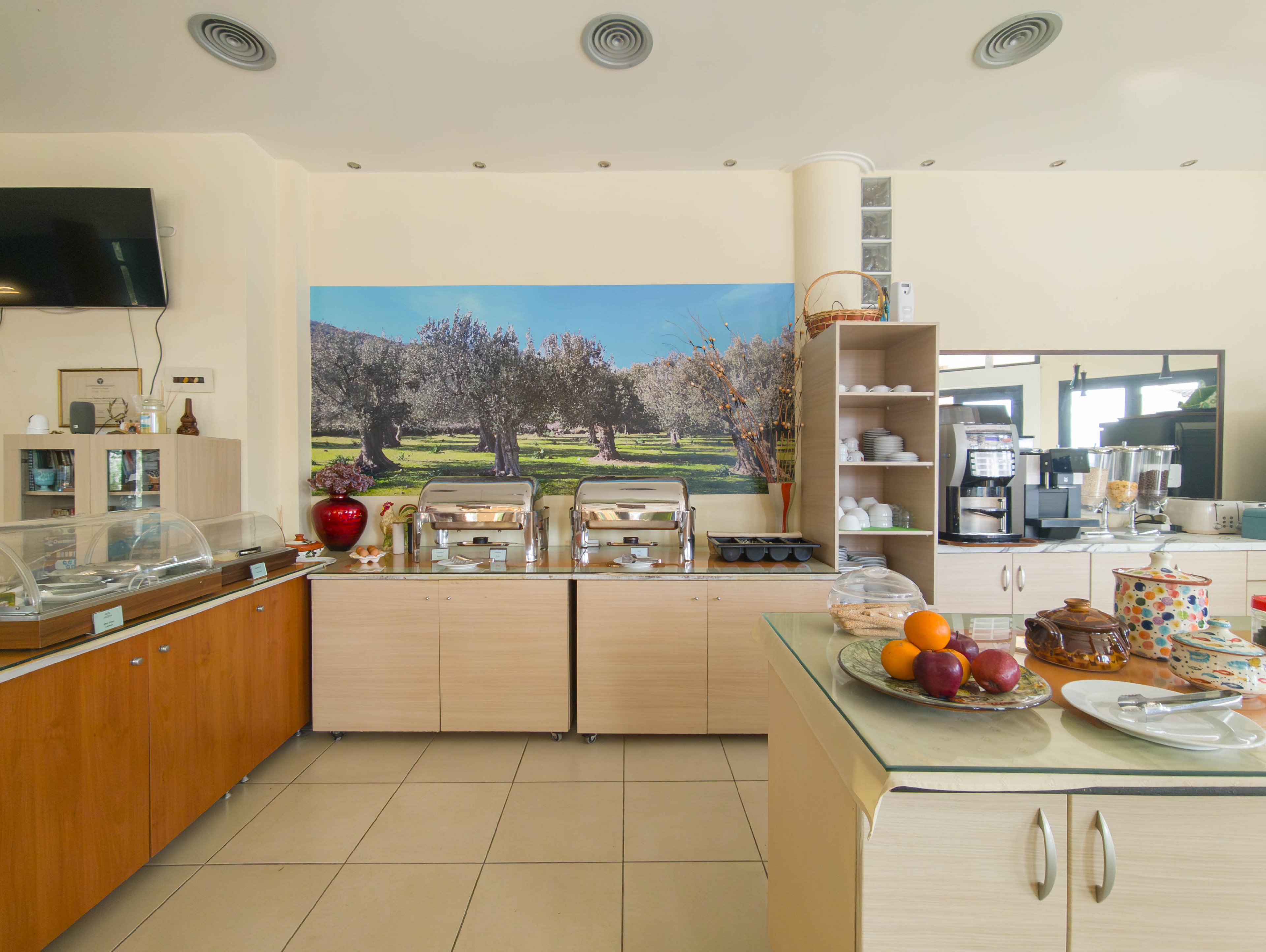 A self-service breakfast buffet is arranged on light wood counters in a bright dining area. Chafing dishes, coffee machines, and stacked plates are ready for guests. In the foreground, a glass-topped island displays fresh fruit and assorted pastries on plates. A large landscape photograph of olive trees decorates the back wall, adding color to the neutral interior. The space appears clean, organized, and inviting for morning meals.
