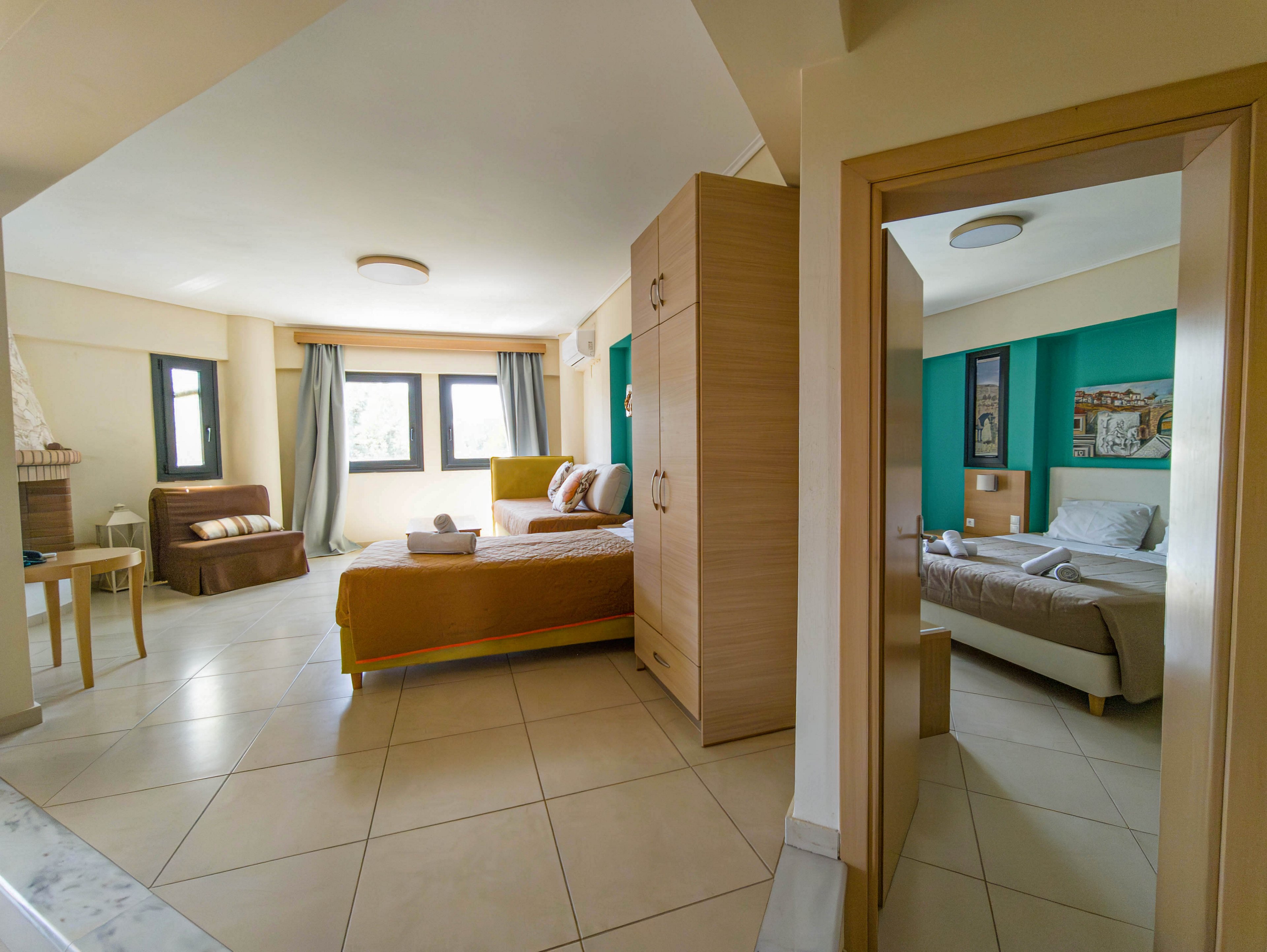 A spacious, contemporary hotel suite features a main bedroom with a double bed, sofa, and large windows with curtains. Neutral tile flooring and light wood furniture create a clean, minimalist look. Through an open doorway, a second bedroom with teal accent wall and framed artwork is visible. The overall atmosphere is bright, comfortable, and inviting, suitable for vacation or short-term stays.