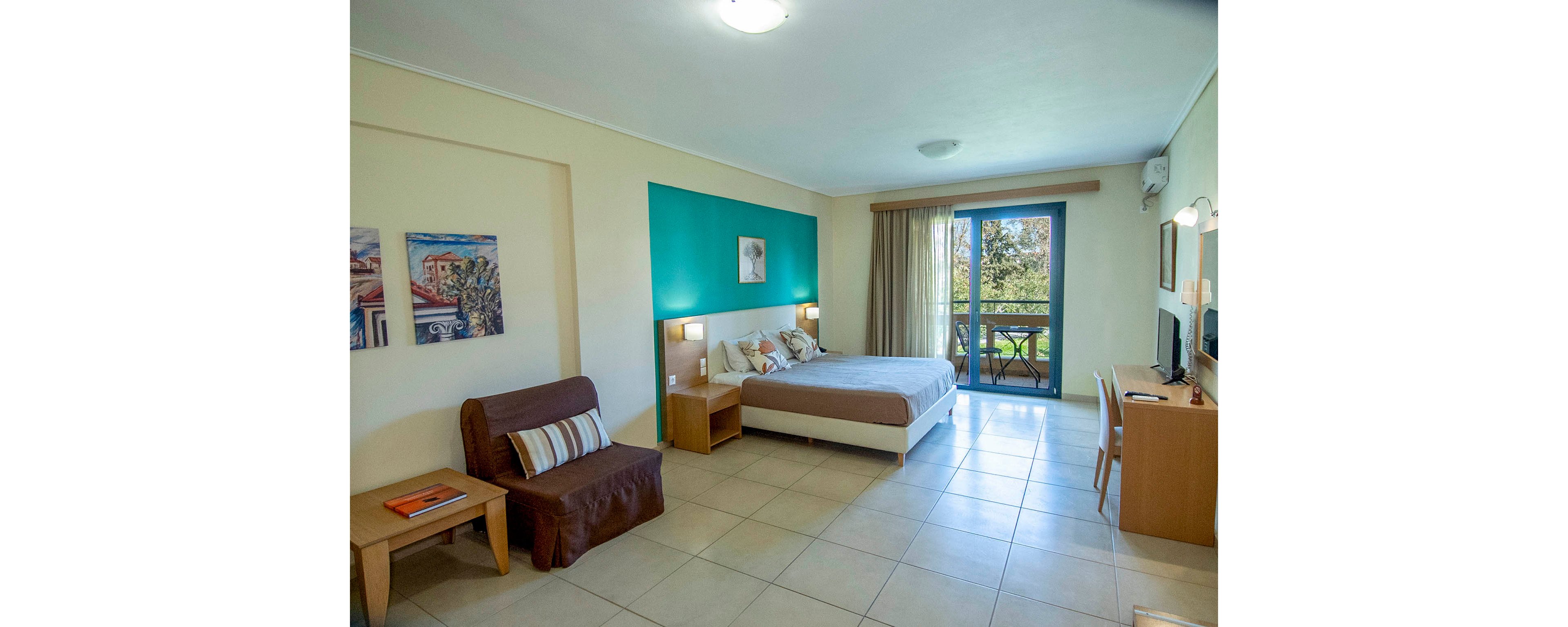 Spacious hotel room featuring a double bed with decorative pillows and a teal accent wall. A small sofa and wooden side table sit to the left, while a desk with chair and mirror occupies the right side. Large glass doors open to a balcony with trees visible outside, allowing natural light to fill the tiled floor space. Wall art and soft lighting create a clean, inviting atmosphere suitable for vacation or business travel.