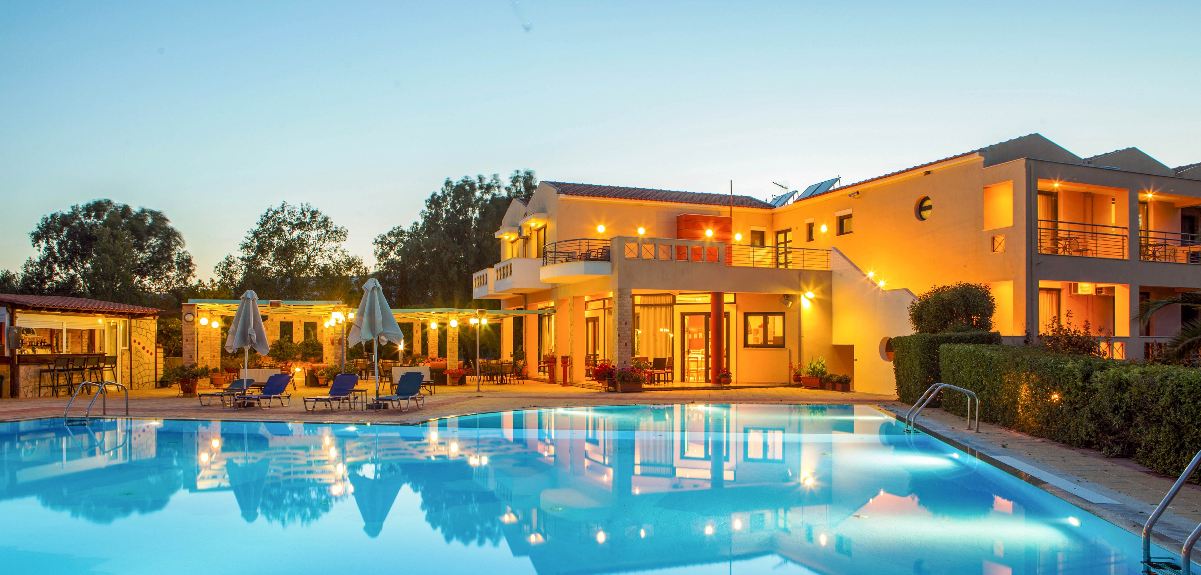 A modern resort hotel is shown at dusk with its exterior warmly lit against the evening sky. In the foreground, a large swimming pool reflects the building lights and surrounding trees. Sun loungers and umbrellas line the poolside, suggesting a relaxing vacation setting. The tranquil atmosphere and soft blue and yellow tones create an inviting, serene mood.