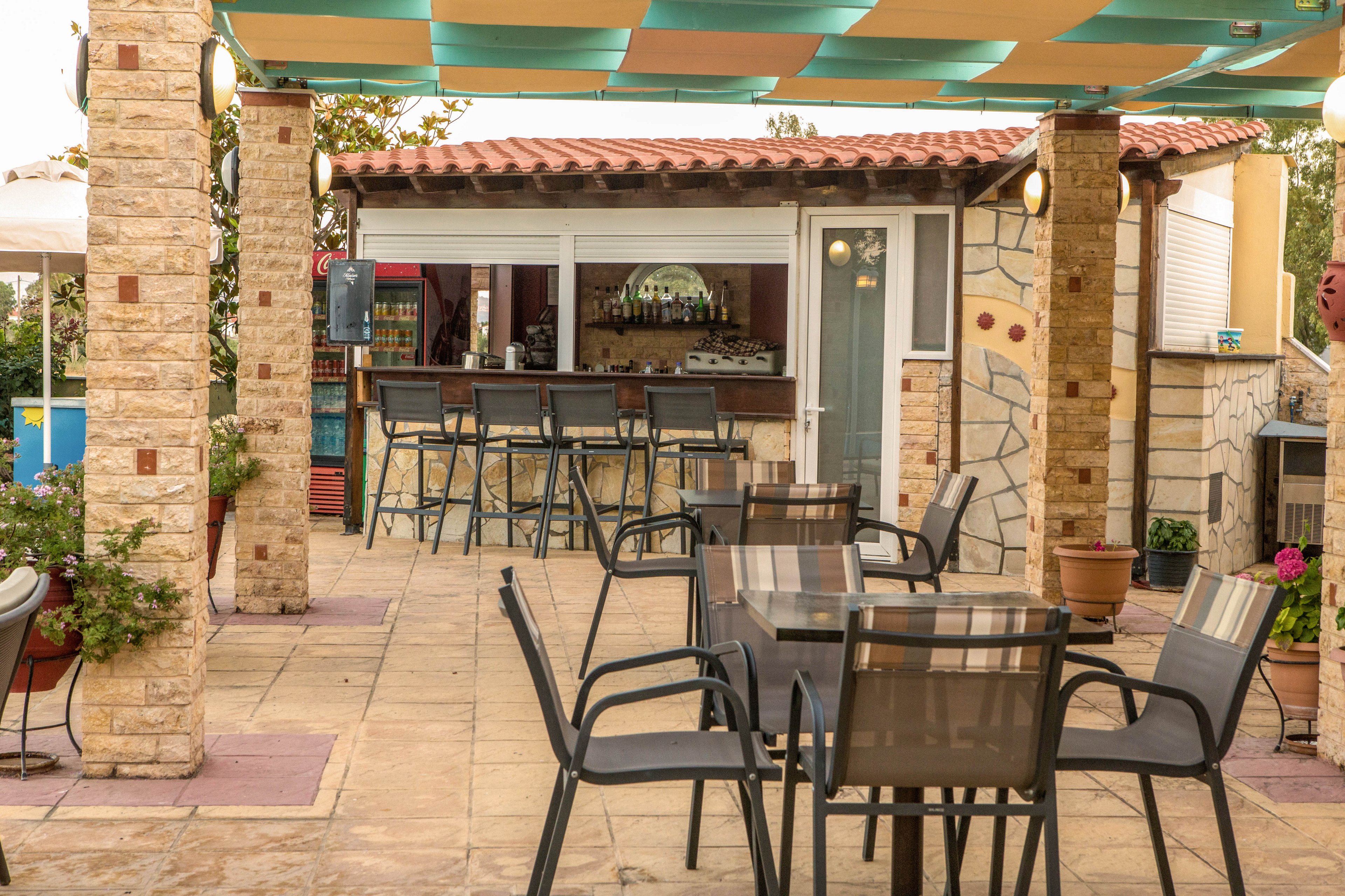 An inviting outdoor bar area features several high stools lined up along a counter in front of a small building. In the foreground, multiple tables with modern chairs are arranged on a tiled patio beneath a colorful fabric canopy. Stone columns and potted plants add a rustic touch to the setting. Warm lighting and soft colors create a relaxed, casual atmosphere suitable for socializing and dining.