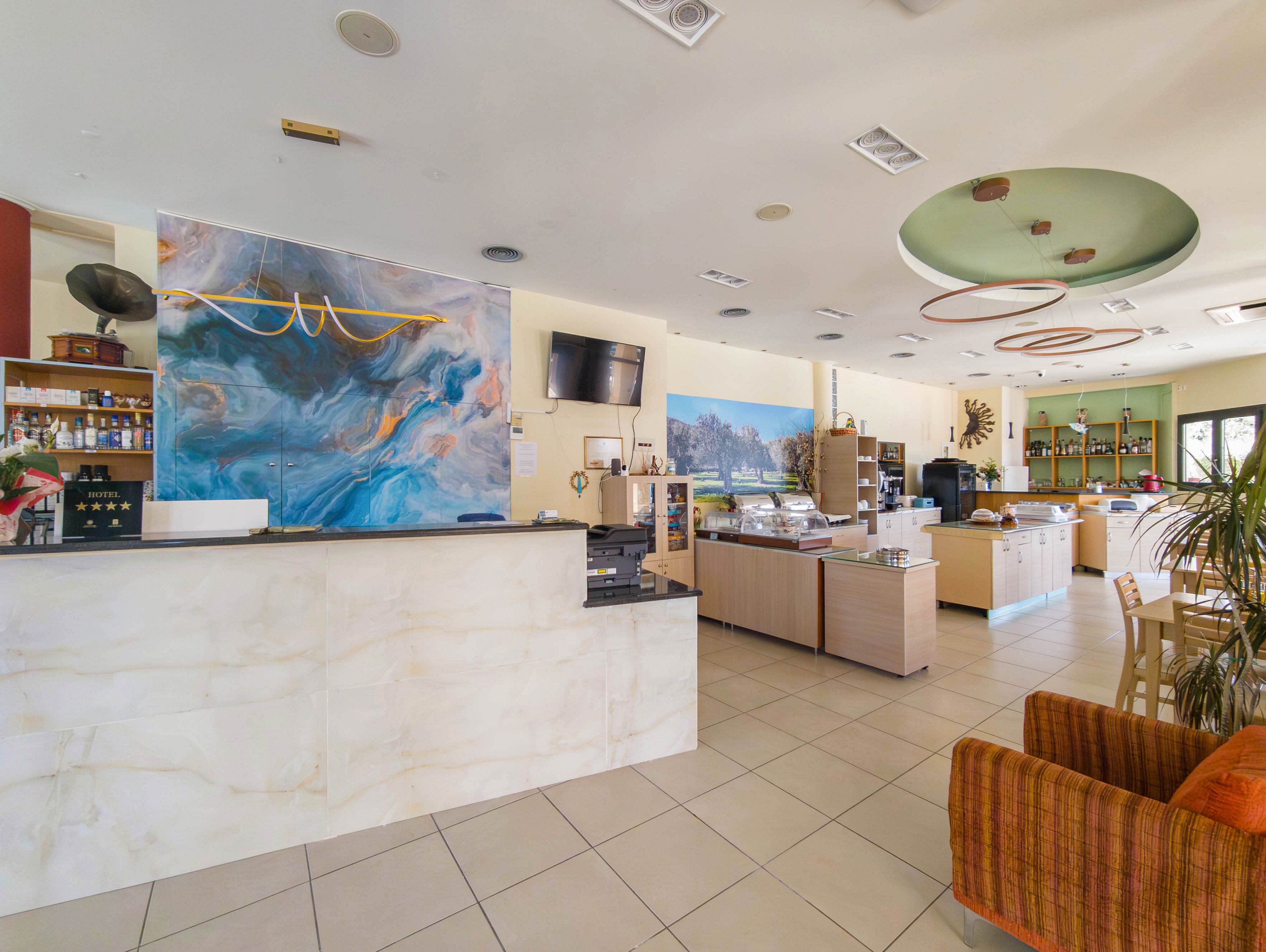 Spacious café interior featuring a large front counter and open service area. The setting includes display cases, coffee machines, and shelves with bottles and jars along the back wall. A colorful abstract mural and circular ceiling cutouts add contemporary visual interest. Natural light enters through windows, illuminating tiled floors and comfortable seating with an orange armchair.
