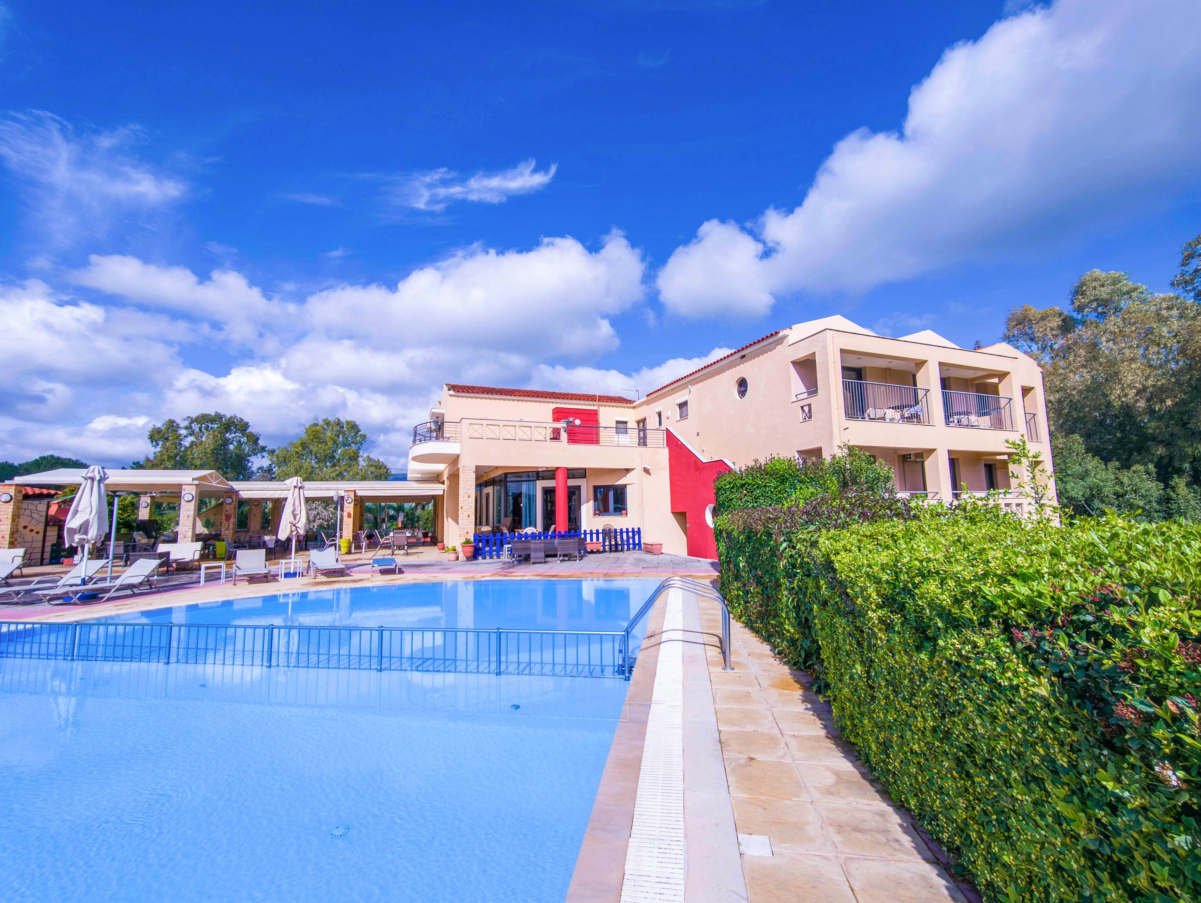 A contemporary villa with cream and red exterior walls overlooks a large outdoor swimming pool. Sun loungers and umbrellas are arranged along the poolside terrace, suggesting a relaxing vacation setting. Lush green hedges border the pool area, and trees frame the property in the background. A bright blue sky with scattered clouds enhances the vibrant, sunny atmosphere.