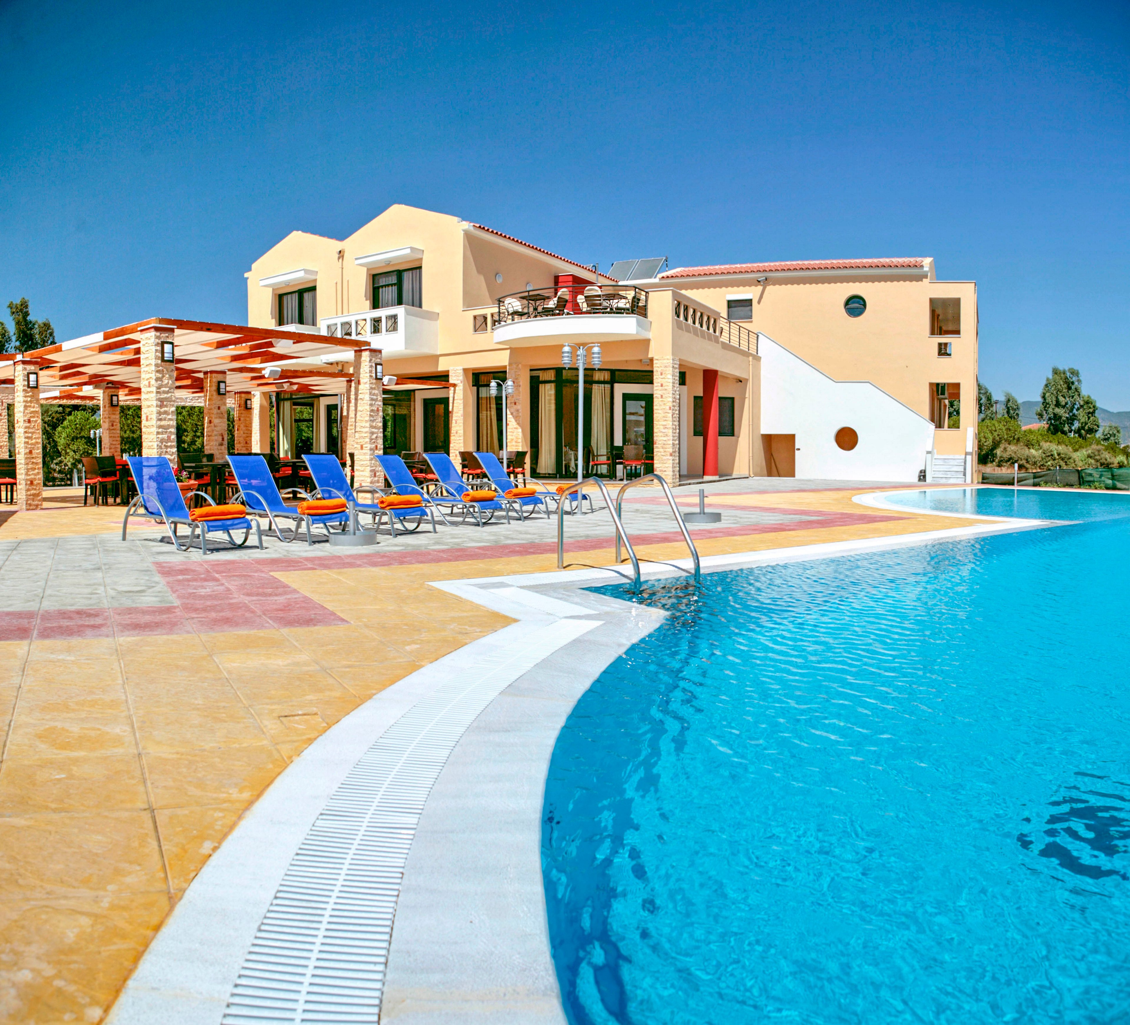 A modern villa-style resort building stands beside a large curved outdoor swimming pool under a clear blue sky. Bright blue lounge chairs are arranged on the colorful tiled pool deck near a wooden pergola. The architecture features warm beige and yellow walls with geometric shapes and balconies. Lush greenery surrounds the property, enhancing the vacation atmosphere.