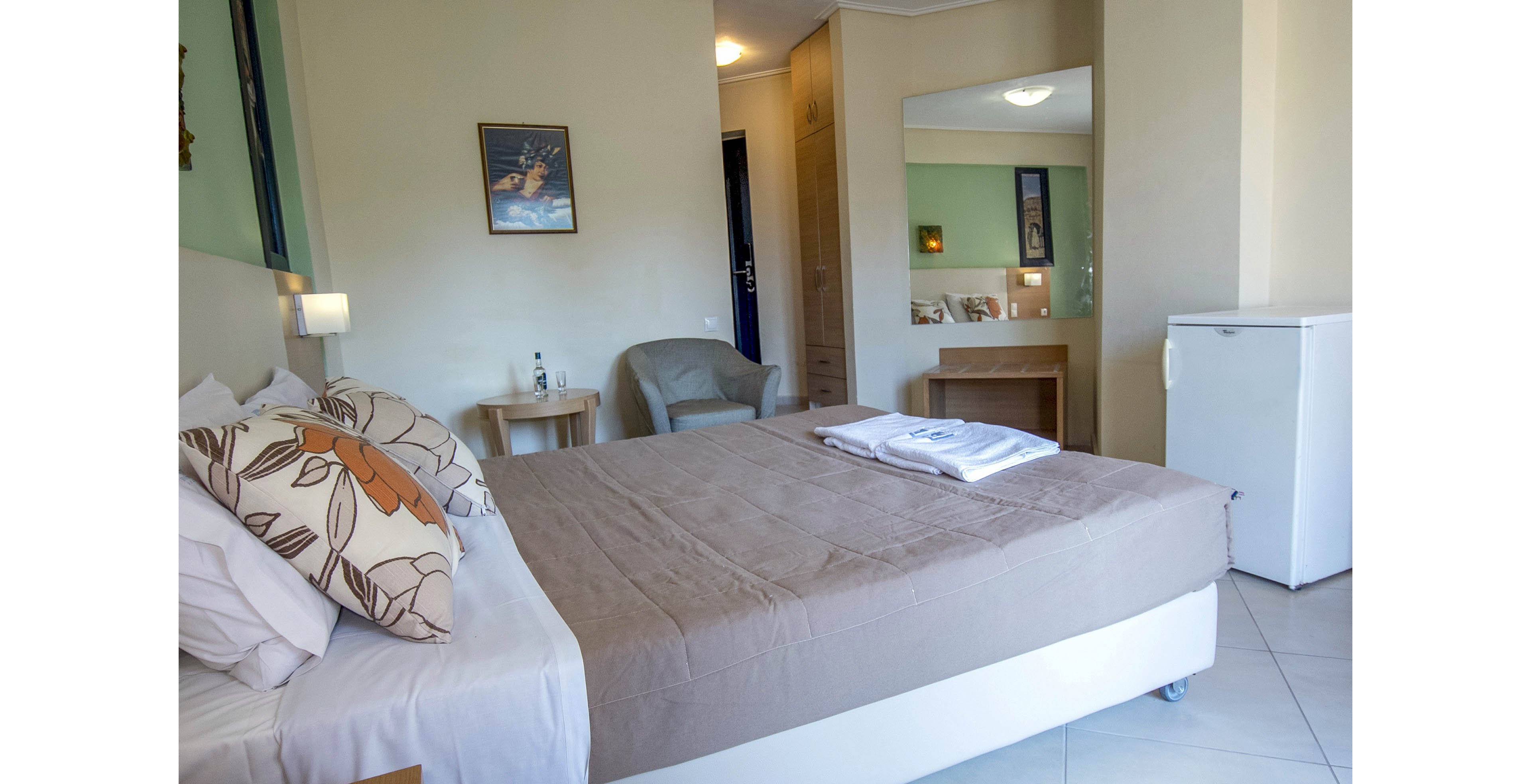 A neatly made double bed with patterned pillows is the focal point of this simple hotel room. The space includes a small table with a chair, a dresser with mirror, and a compact white refrigerator. Neutral walls and soft lighting create a calm, welcoming atmosphere. Folded towels are placed on the bed, suggesting readiness for guests.
