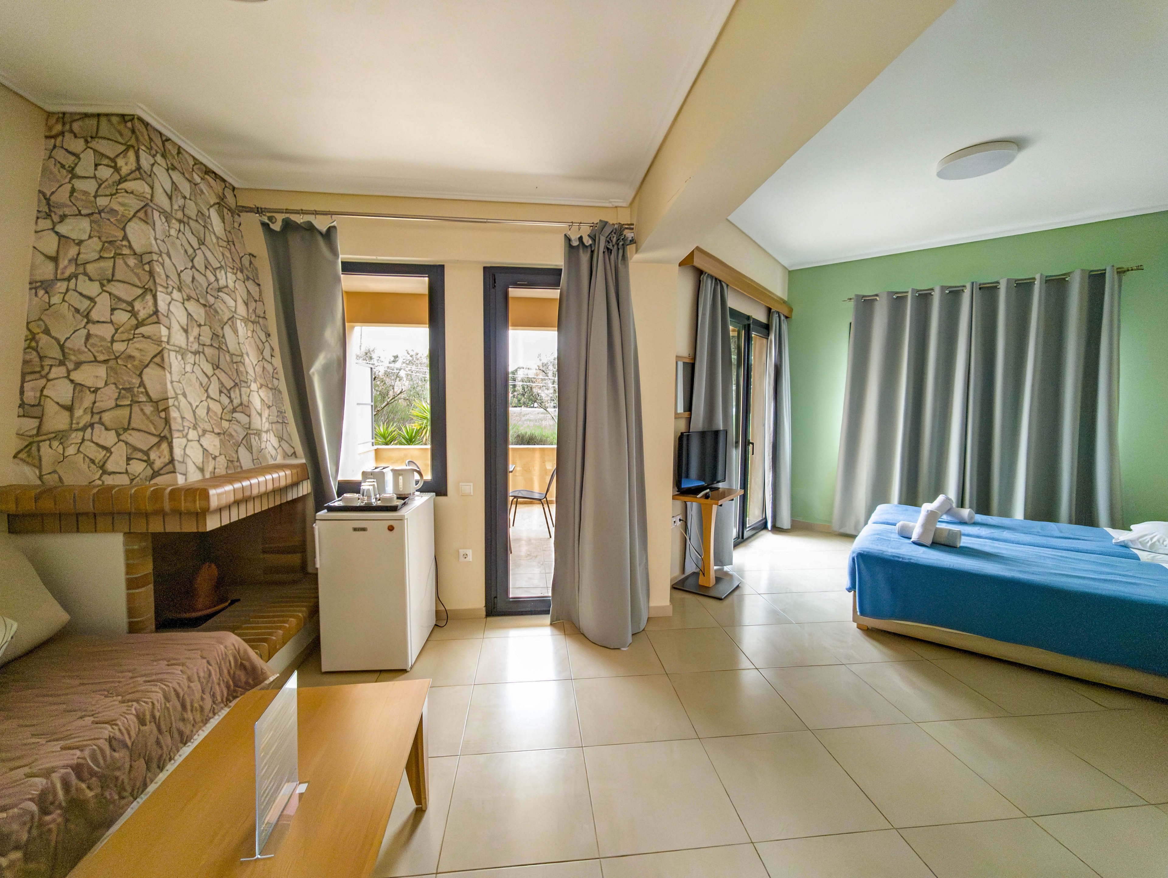 A spacious hotel room features a neatly made double bed with blue bedding and towels placed on top. Large windows and a glass door open to a balcony, allowing natural light to fill the space. The room includes a small refrigerator, electric kettle, and a television on a stand, with neutral tile flooring and light-colored curtains. A stone-faced fireplace and simple wooden furniture add a cozy touch to the modern interior.