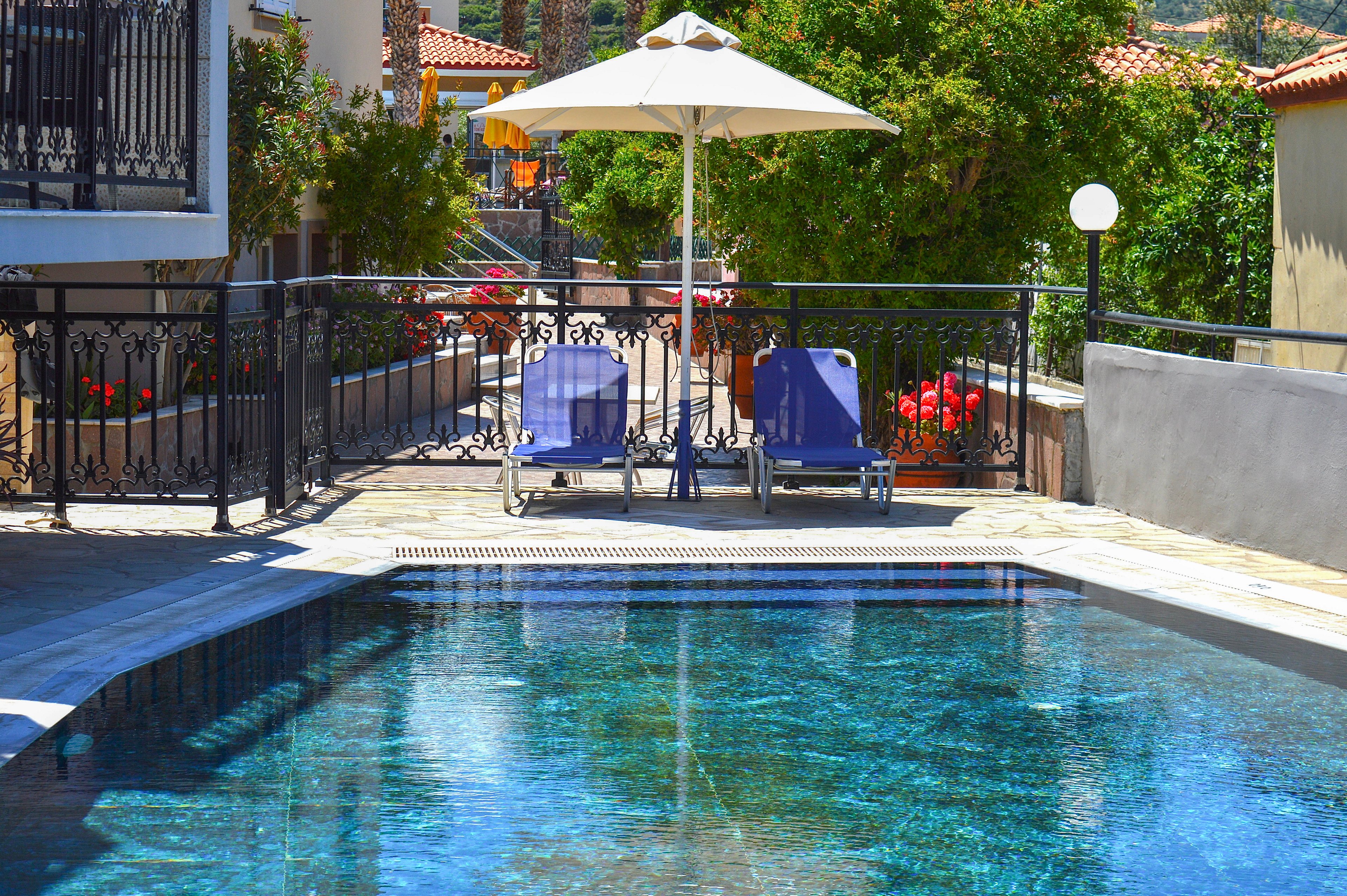 A clear blue swimming pool reflects sunlight in a cozy outdoor courtyard. Two empty lounge chairs sit beneath a large white umbrella beside the pool. Decorative black railings, flowering plants, and red-tiled roofs suggest a Mediterranean-style hotel or villa setting. The scene feels quiet and inviting, ideal for relaxation on a warm summer day.