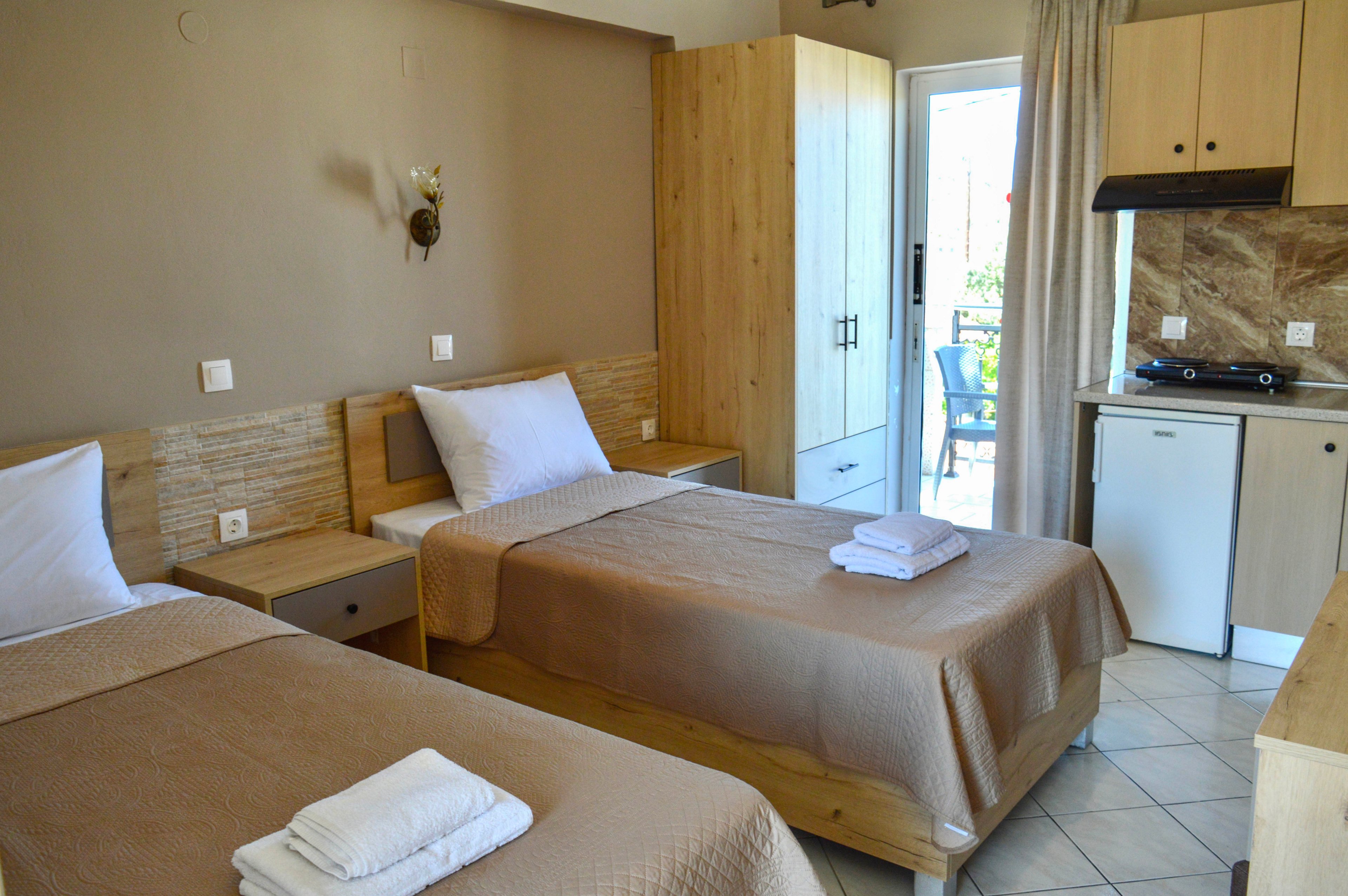 The image shows a compact hotel room with two single beds neatly made with beige bedspreads and white towels. A small kitchenette with a mini fridge, cooktop, and light wood cabinets is visible on the right side. Neutral walls, tiled flooring, and matching wooden furniture create a warm, simple interior. A glass door at the back opens to a balcony, allowing natural light into the room.