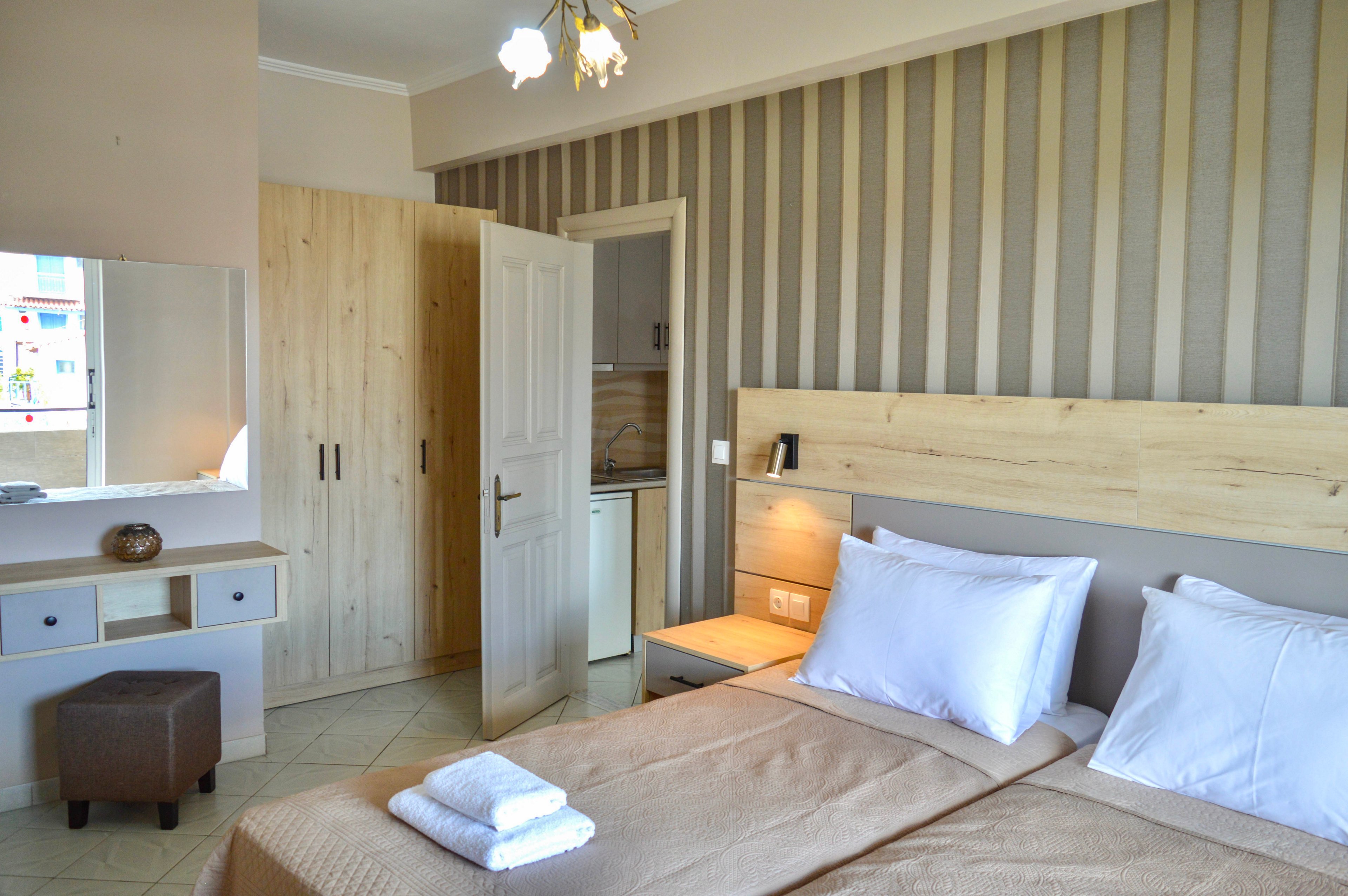 A neatly made double bed with folded white towels sits in a modern hotel bedroom. The room features light wood furniture, including a wardrobe, headboard, and dressing table with mirror. A small kitchenette is visible through an open door, adding convenience for guests. Soft lighting and beige tones create a calm, welcoming atmosphere suitable for travel or vacation stays.