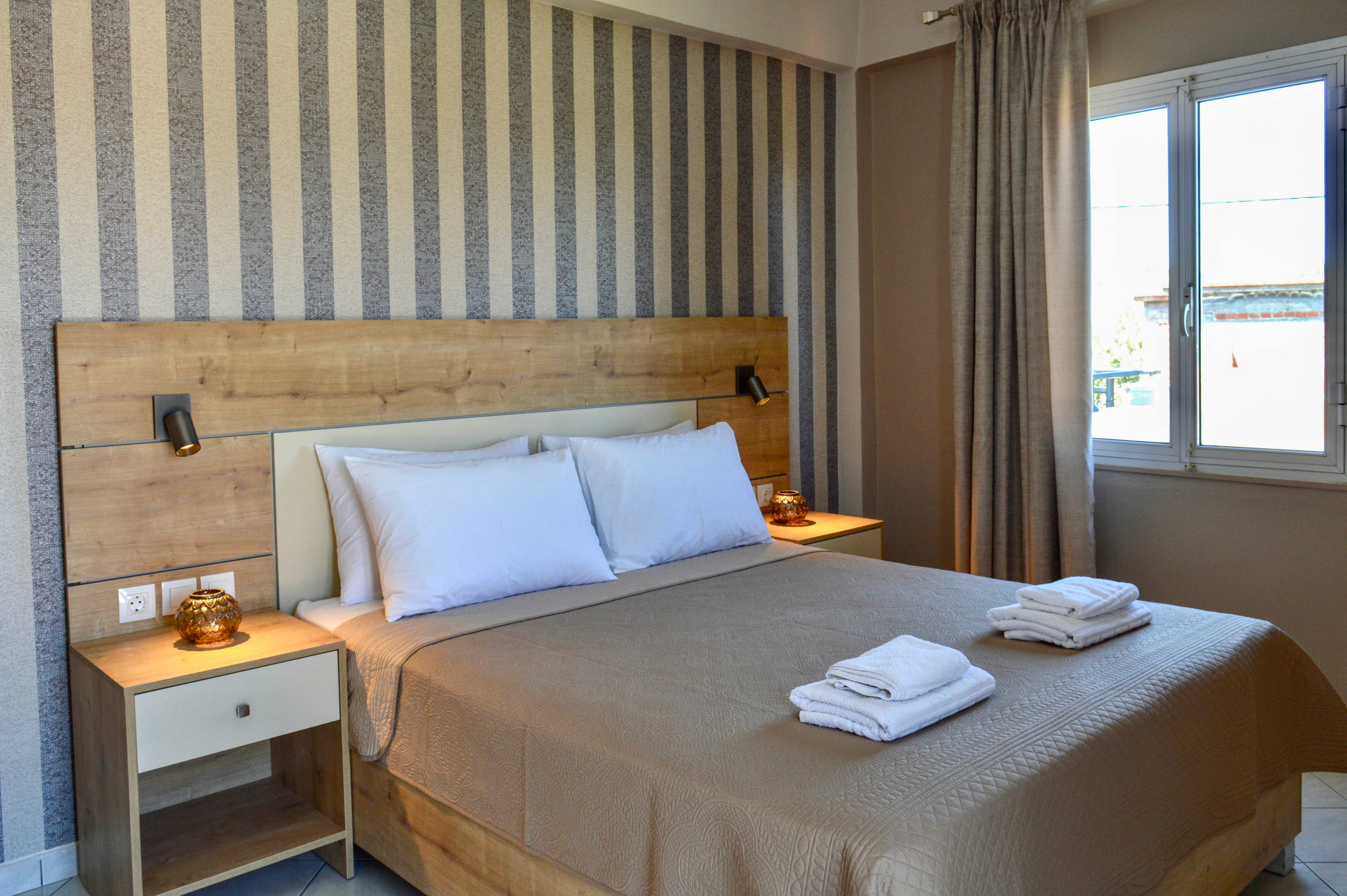 A neatly made double bed with beige bedding stands against a wooden headboard in a modern bedroom. The room features striped wallpaper, matching bedside tables, and small wall-mounted reading lights. Folded white towels are placed on the bed, suggesting a hotel or guest accommodation. Natural light enters through a large window with neutral curtains, creating a calm and inviting atmosphere.
