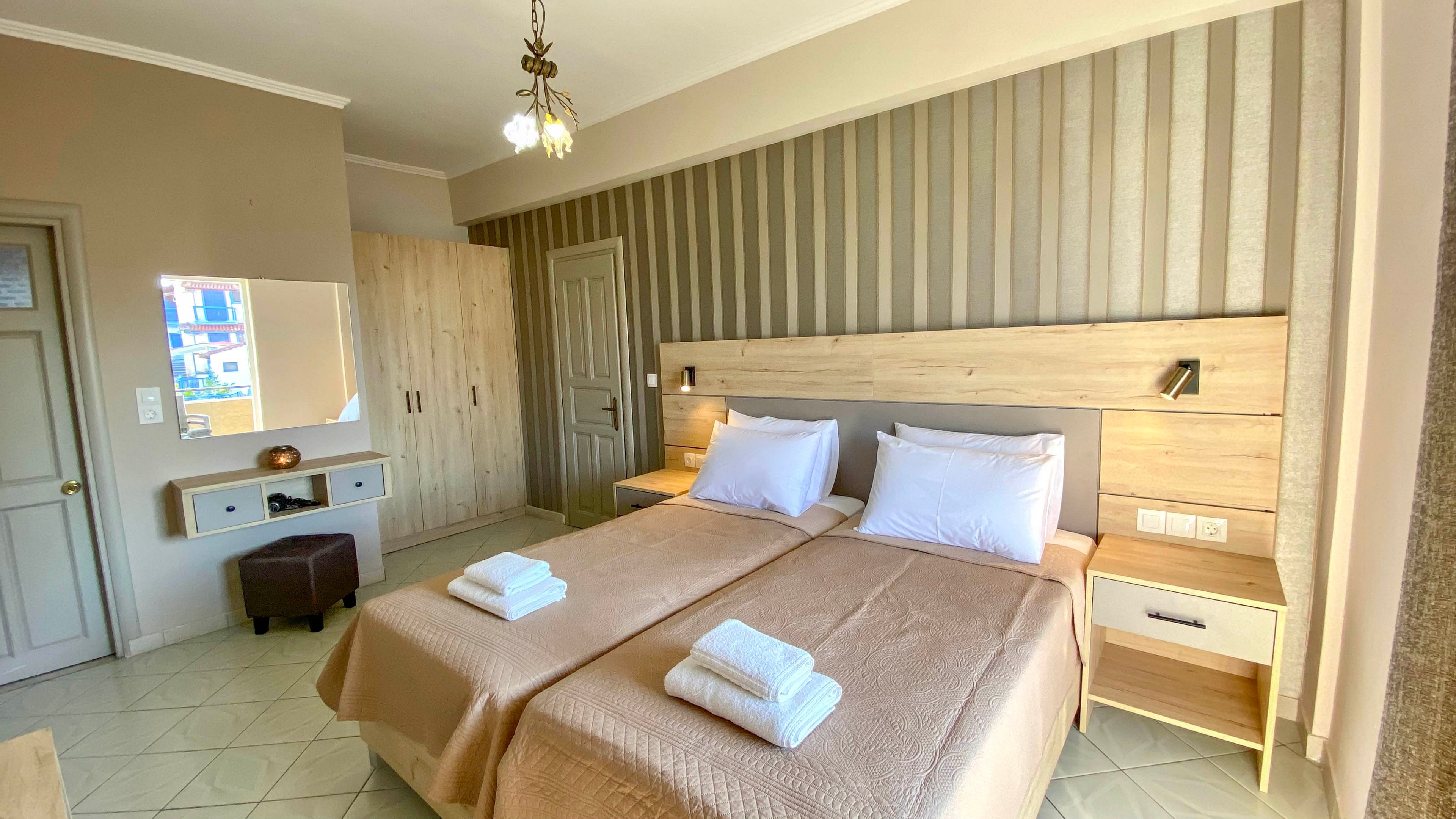 Well-lit hotel room featuring two neatly made twin beds with beige bedspreads and stacked white towels. The space includes light wood bedside tables, a wardrobe, and a dressing table with a mirror and stool. Vertical striped wallpaper and neutral wall colors create a modern yet cozy atmosphere. Natural light enters from a window out of frame, highlighting the clean and organized interior.