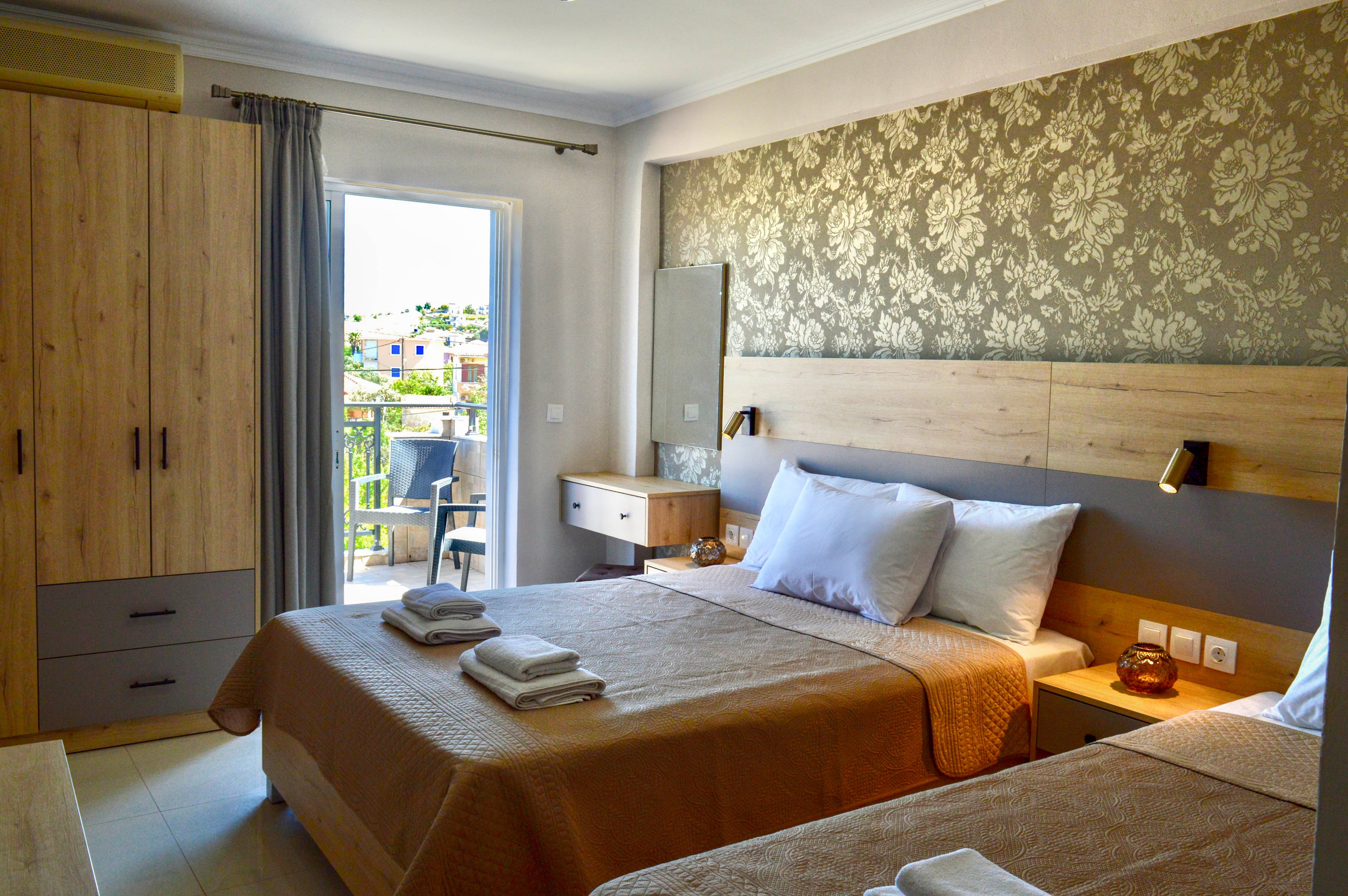 A neatly arranged hotel bedroom features two beds with beige bedspreads and white pillows. Warm wooden furniture, including a wardrobe, bedside tables, and a small desk, complement the neutral decor. Natural light streams in through a glass door that opens onto a balcony with outdoor seating and a view of nearby buildings. Wall-mounted reading lights and folded towels on the beds add to the welcoming, comfortable atmosphere.