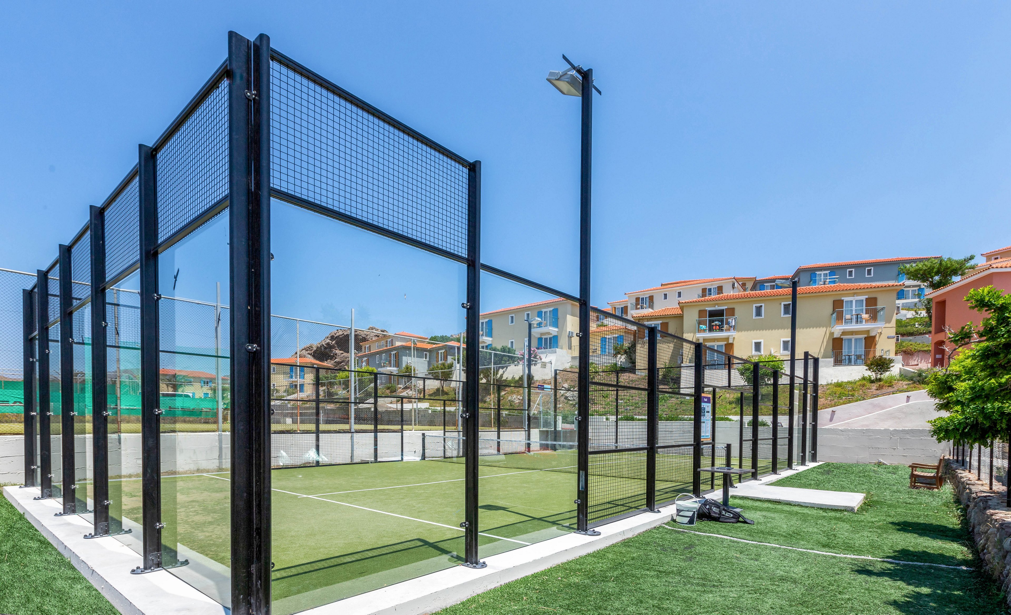 An empty outdoor padel court is enclosed by tall black metal and mesh fencing under a clear blue sky. The court surface is green with white lines, surrounded by artificial turf and a paved walkway. In the background, multi-story residential buildings with colorful facades overlook the sports area. Sports bags and equipment rest near the court, suggesting recent or upcoming play.