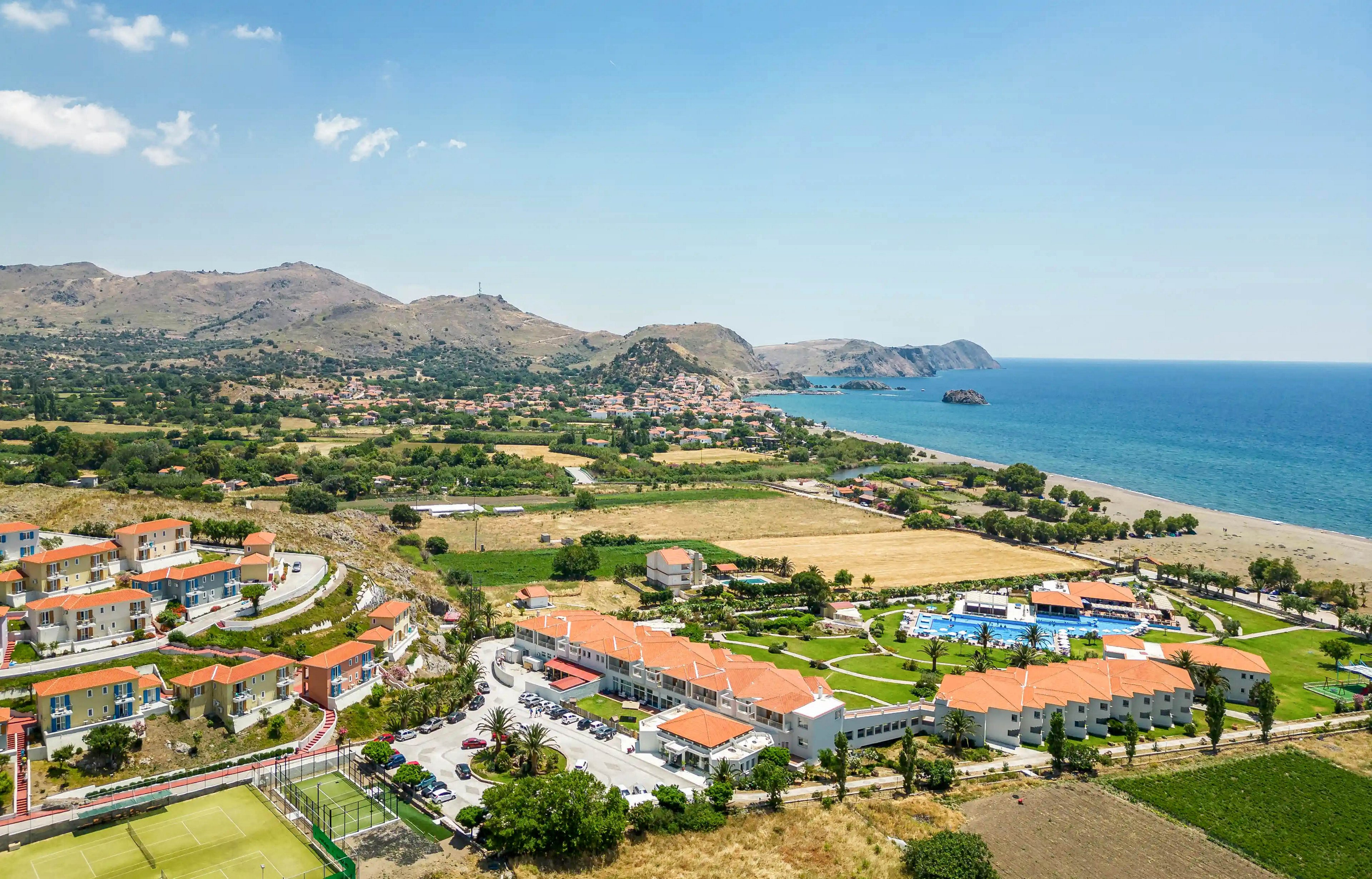 Aerial view of a coastal resort complex with orange-roofed buildings near the shoreline. The scene includes swimming pools, sports courts, and parking areas surrounded by fields and greenery. Rolling hills and scattered houses extend into the distance, meeting the blue sea on the right. The overall atmosphere is sunny and relaxed, suggesting a vacation destination.