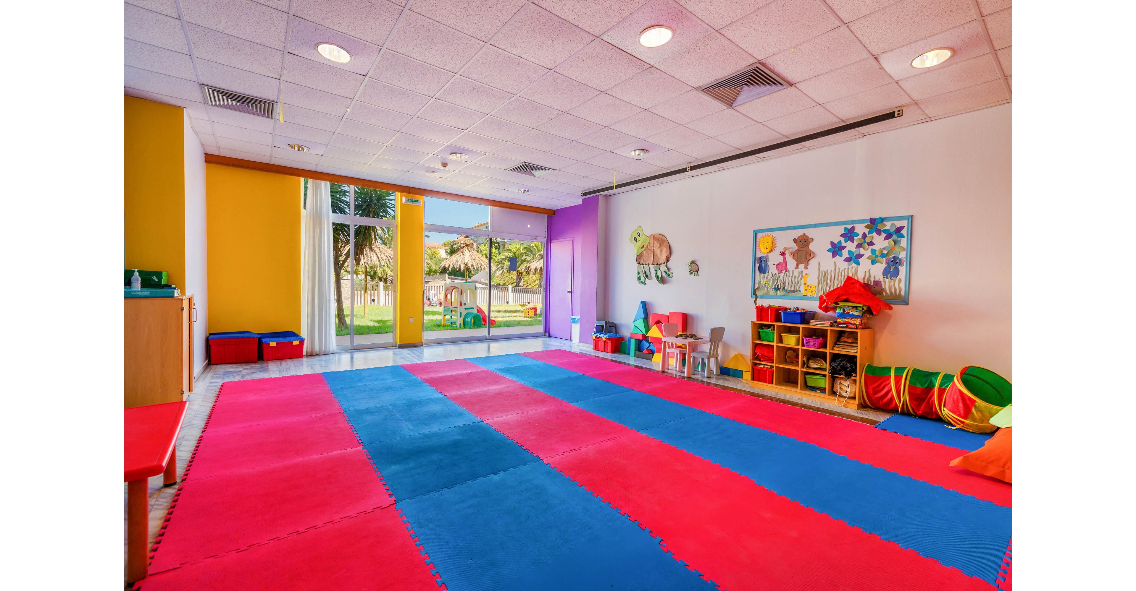 Bright, spacious playroom featuring large red and blue foam floor mats. The room has colorful walls and large windows overlooking a green outdoor area with palm trees. Shelves hold toys, balls, and art supplies, and a bulletin board displays children’s artwork. The overall atmosphere is cheerful and inviting, suitable for kids’ activities and early learning.