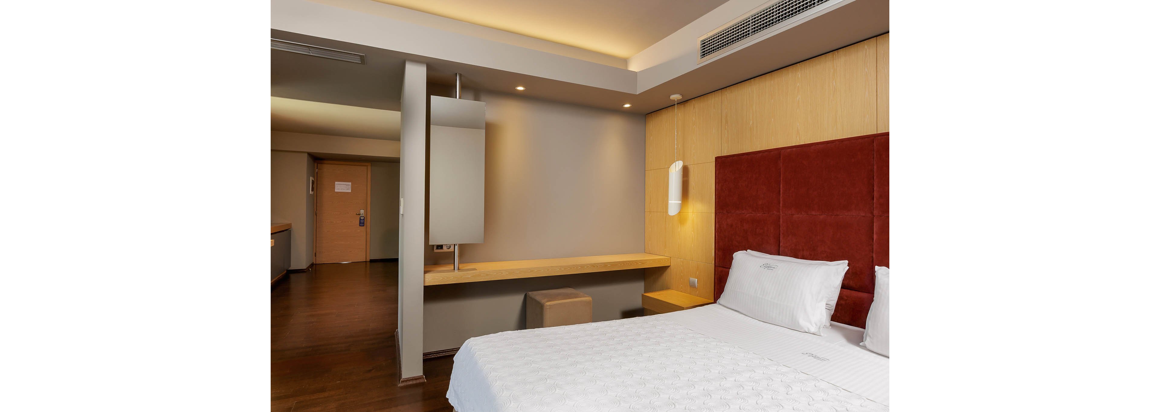 A contemporary hotel room features a neatly made double bed with white linens and a tall upholstered headboard. The space includes a long wooden desk with a mirror and a small ottoman beneath it. Warm recessed lighting and wall-mounted bedside lamps create a cozy atmosphere. The room has clean lines, neutral colors, and a simple, uncluttered layout leading toward the entrance door.