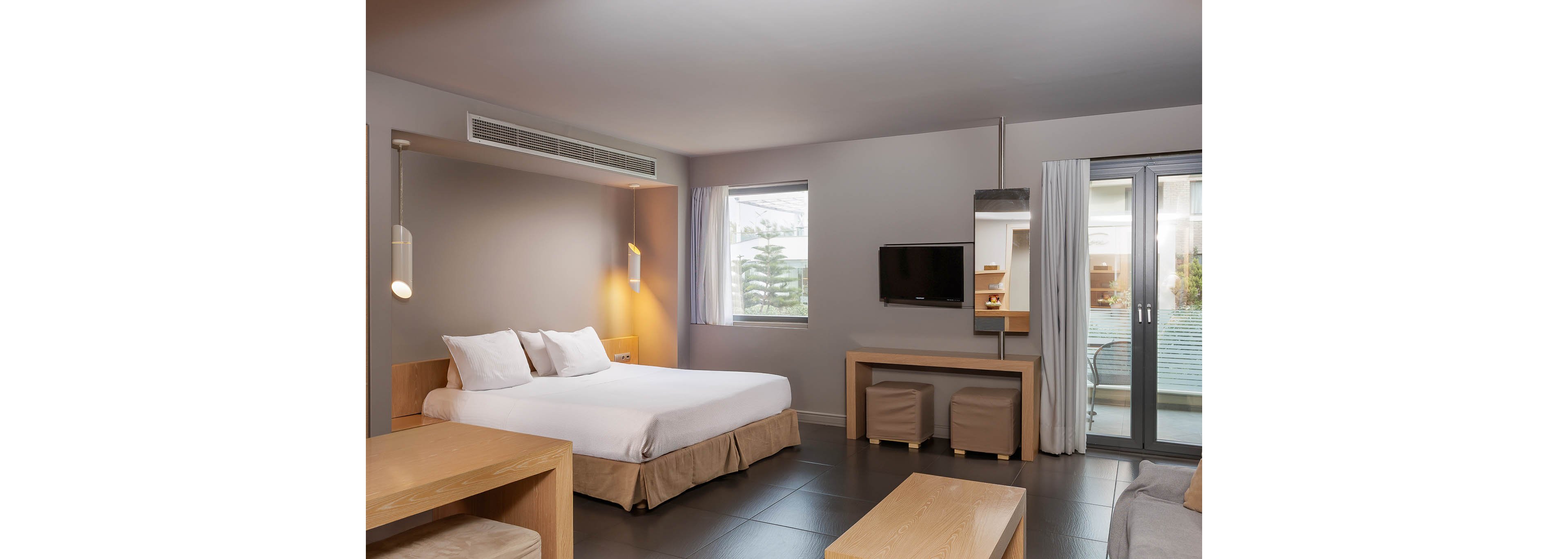 Spacious modern hotel room featuring a large double bed with white linens. The room includes light wood furniture, a wall-mounted flat-screen television, and two upholstered stools beneath a console table. A window and glass doors with white curtains provide natural light and access to a balcony or terrace. Neutral colors and clean lines create a calm, contemporary atmosphere.