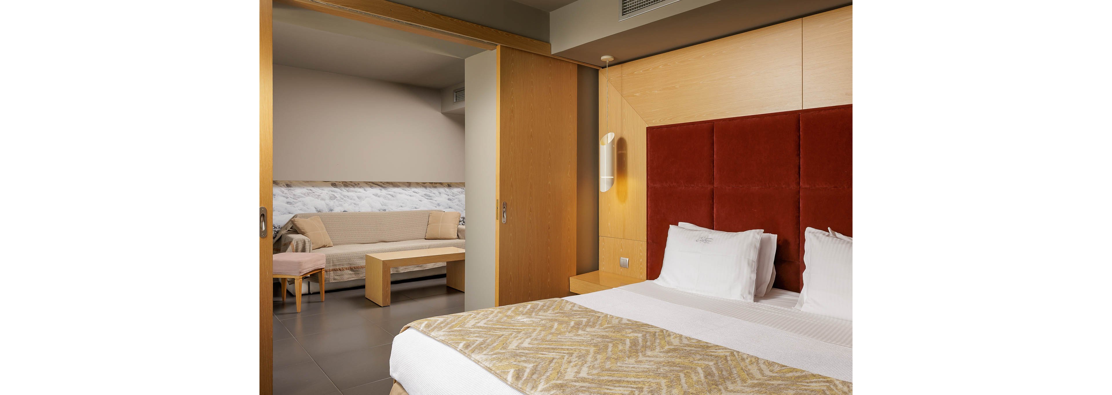Contemporary hotel room interior featuring a neatly made double bed with white linens and patterned blanket. A sliding wooden partition opens to an adjoining sitting area with a sofa, coffee table, and small side table. Warm wood tones, red upholstered headboard, and neutral walls create a calm, minimalist atmosphere. Soft lighting and clean lines emphasize comfort and modern design.