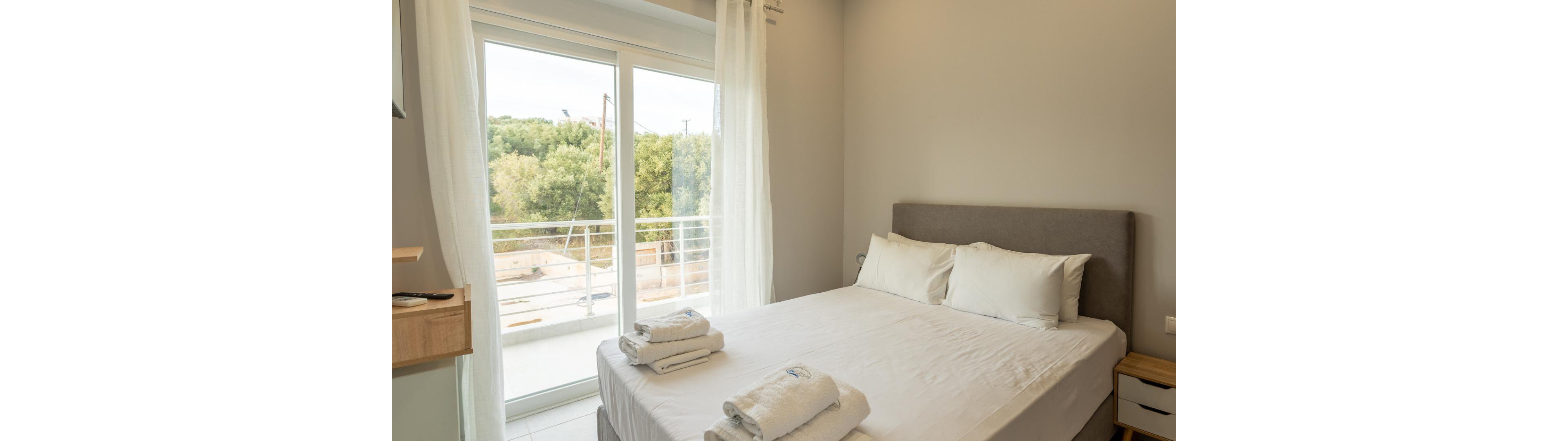 A neatly made double bed with white linens and pillows is the main focus of this modern bedroom. The room features large glass doors that open to a balcony with a view of greenery outside. Freshly folded towels are placed on the bed, and soft natural light fills the space, creating a calm and inviting atmosphere. The walls are painted a light neutral color, enhancing the airy feel.