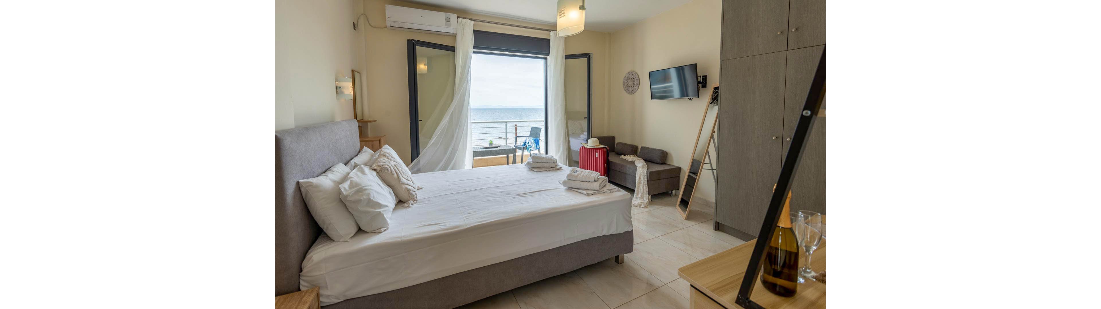 A contemporary hotel room features a large bed with white linens, a wardrobe, and a wall-mounted television. The room opens to a balcony overlooking the ocean, with sheer curtains blowing gently. Key visuals include a red suitcase, a bottle of wine with glasses, and folded towels on the bed, creating a welcoming and relaxed atmosphere.