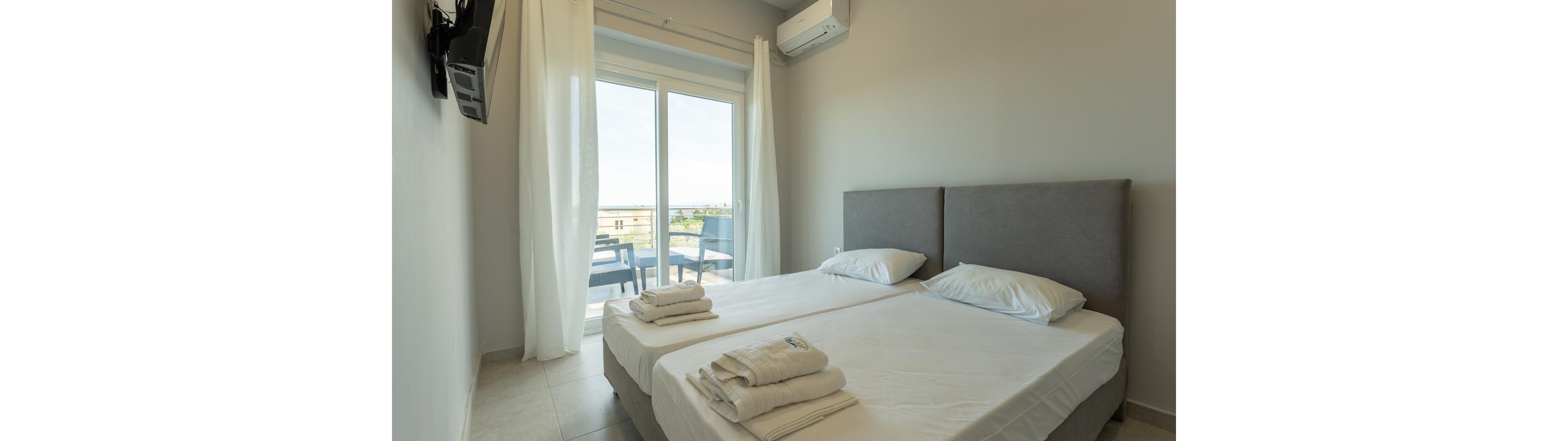 A modern hotel room features two twin beds with gray headboards, neatly arranged with folded towels on each bed. The space is filled with natural light from large glass doors leading to a balcony with outdoor seating. The room has a minimalist style with neutral tones, white curtains, and a wall-mounted air conditioner. A flat-screen TV is visible on the wall, and the overall atmosphere is clean and inviting.